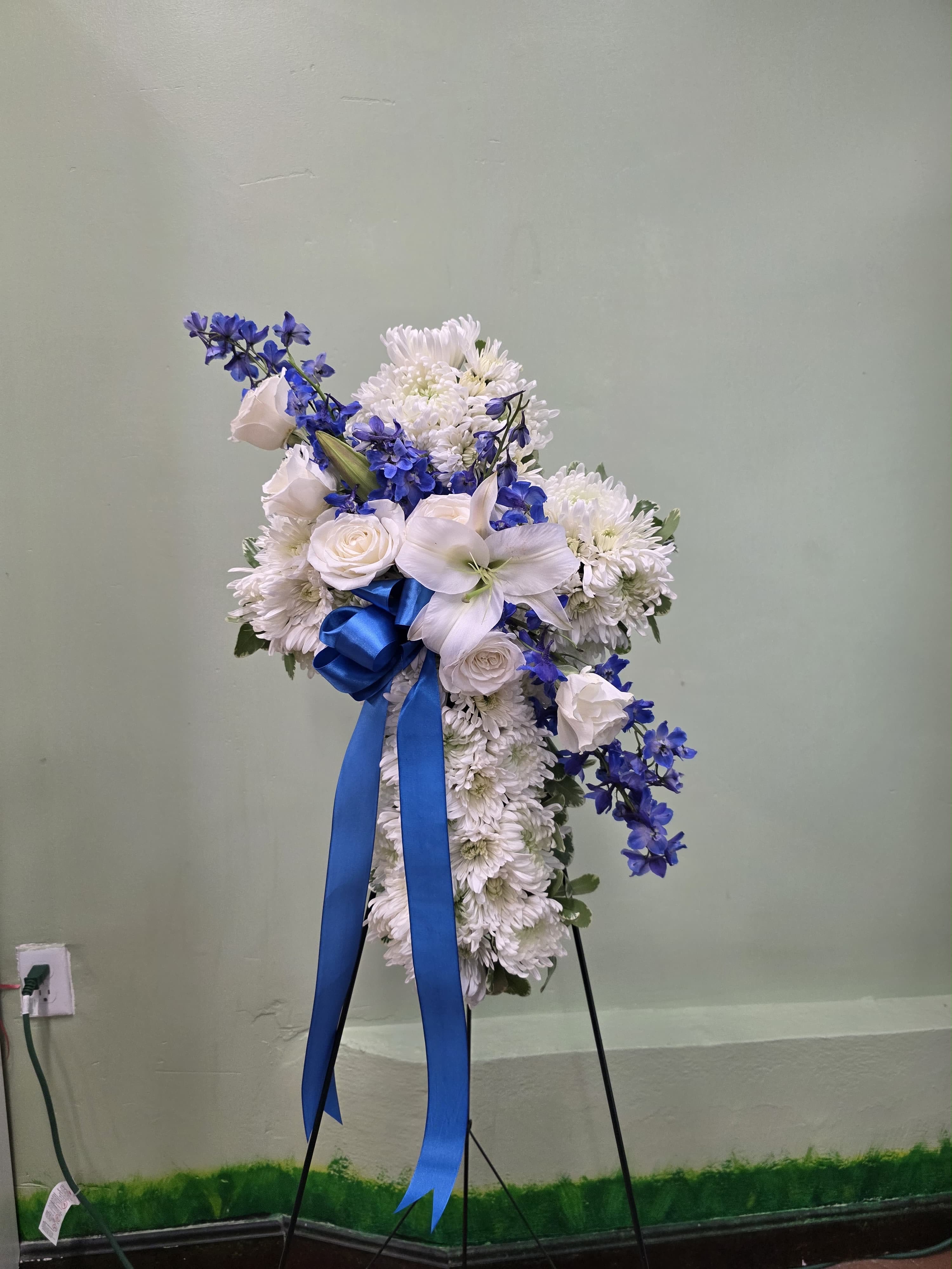 Blue Skies Ahead  - Vivid blue delphinium threads through a cloud of white roses, lilies, and chrysanthemums in this tall standing spray designed for sympathy and memorial services. The vertical, layered silhouette on a standing easel and the large royal-blue ribbon create a formal, respectful presentation that communicates heartfelt condolence to the person you're honoring.