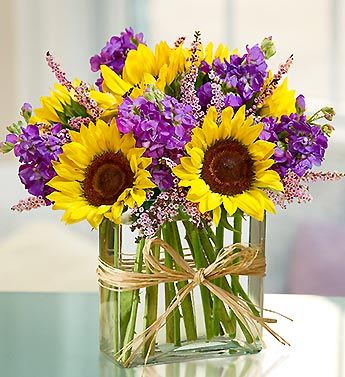 Sunflowers and purple flowers in a clear glass vase with a raffia bow