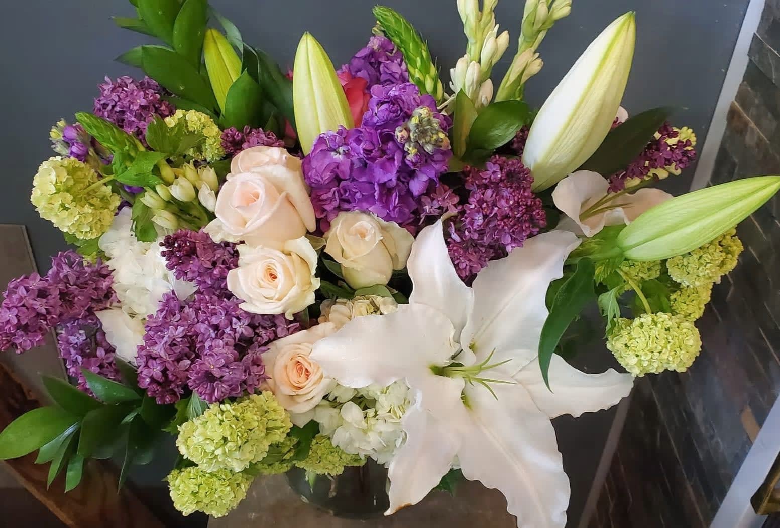 Mixed arrangement of purple lilacs, white lilies, and pale roses in a clear glass vase