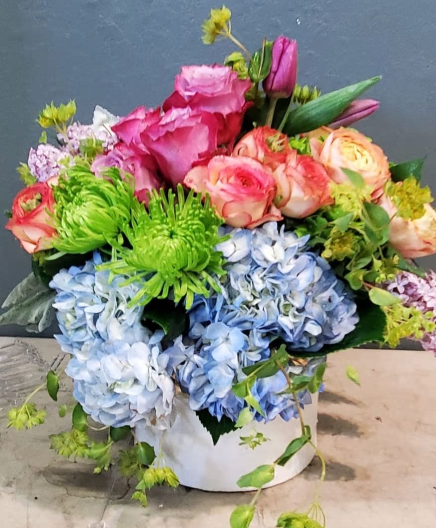 Colorful arrangement of pink roses, green mums, and blue hydrangeas in a white cylinder vase.