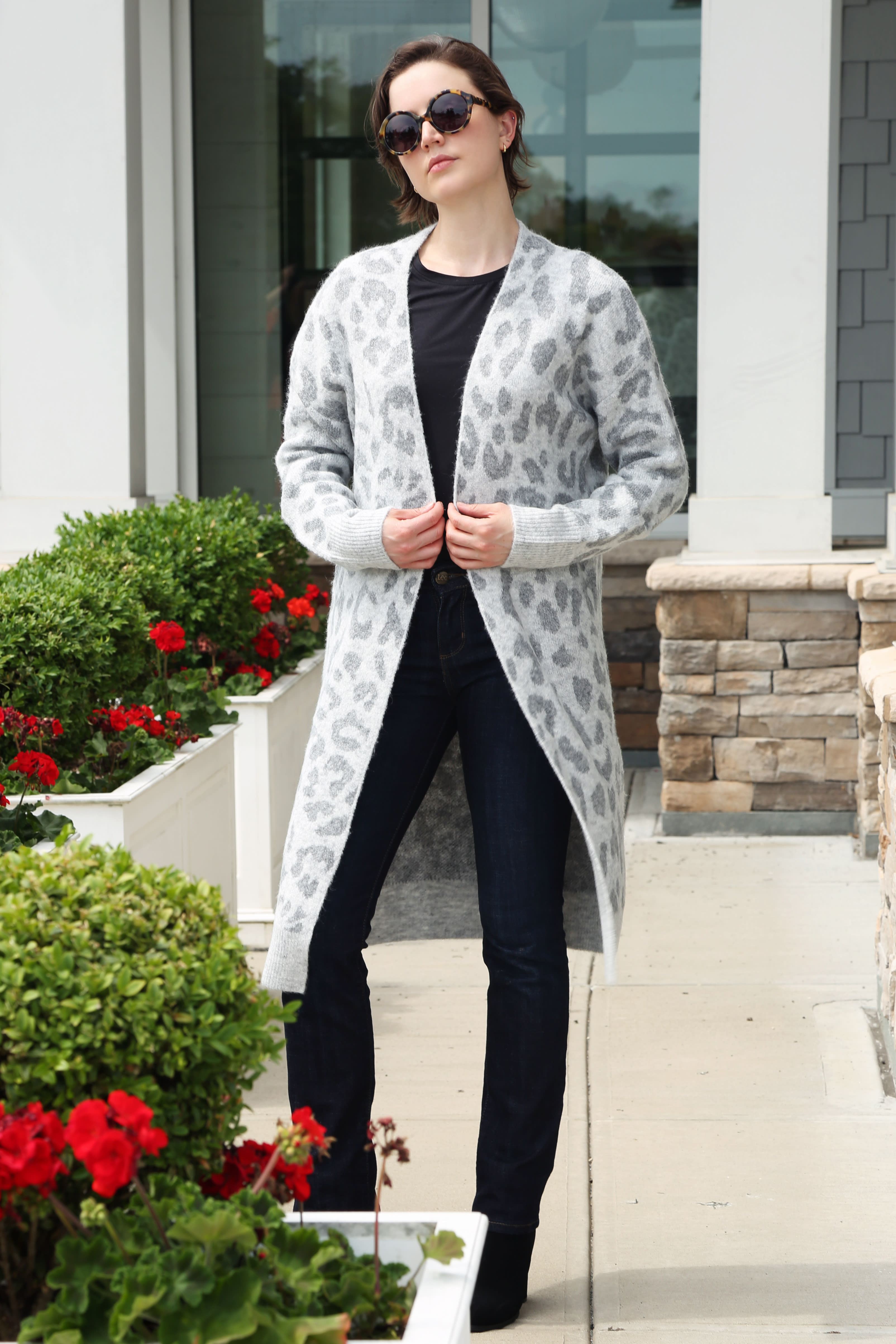 Woman in a long gray patterned cardigan and dark jeans standing by white planters with red flowers
