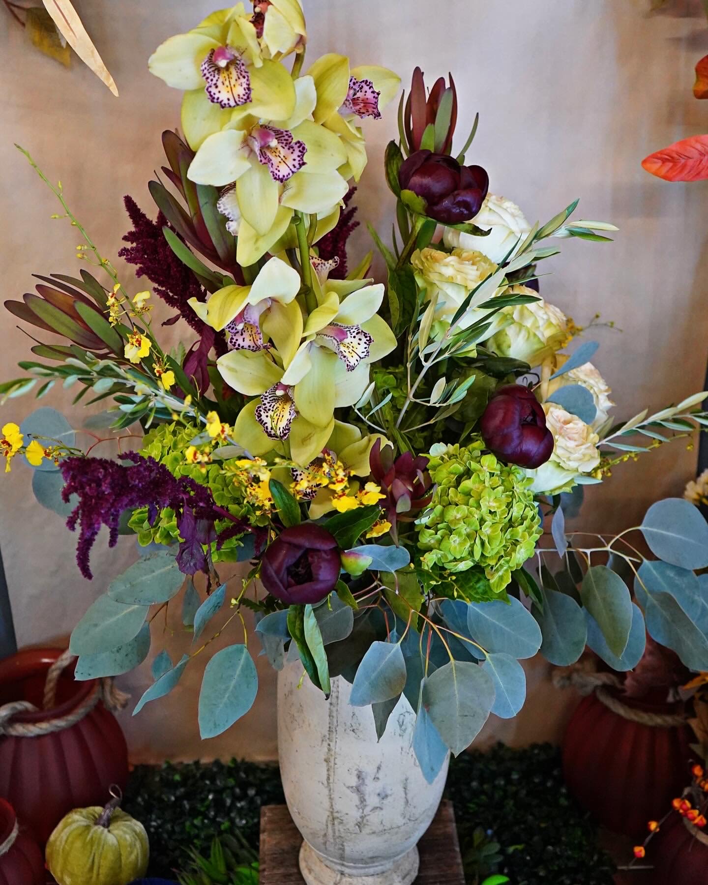 Tall arrangement of green orchids, hydrangea, and dark purple blooms in a white urn-style vase