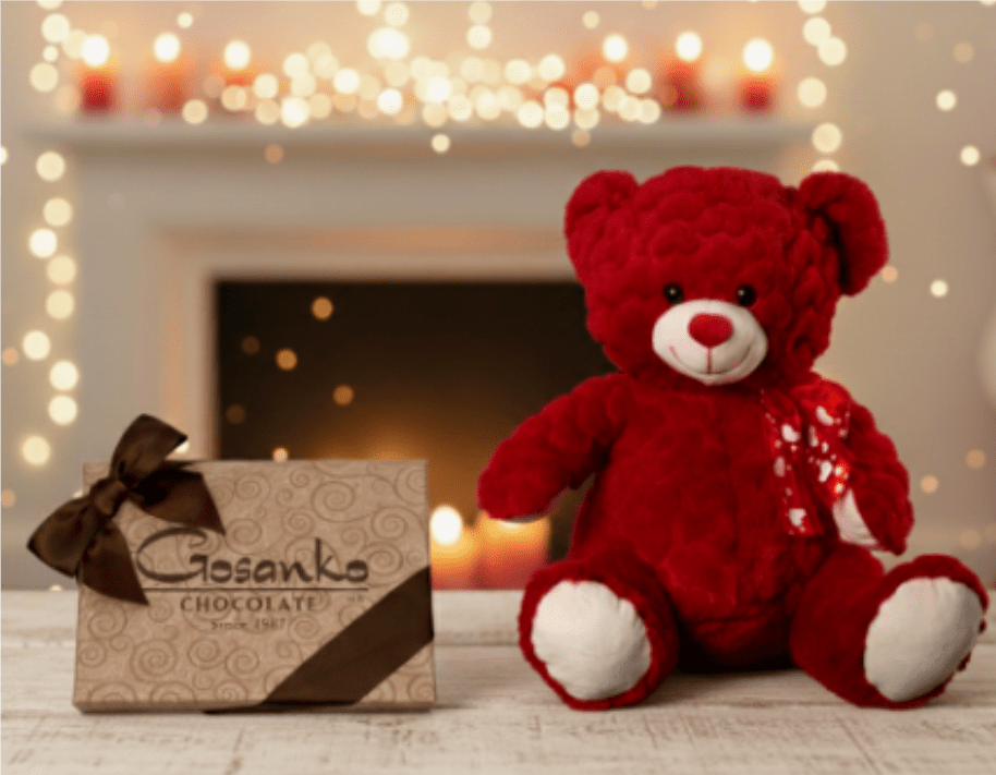 Red teddy bear and decorative chocolate box on a wooden surface with soft candlelight in the background.
