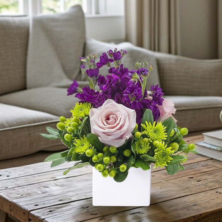 Anna Marie - Anna Marie is a sweet pop of purple with lovely lavender roses, purple stock, and bright green accents in a white cube container appropriate for a coffee table.