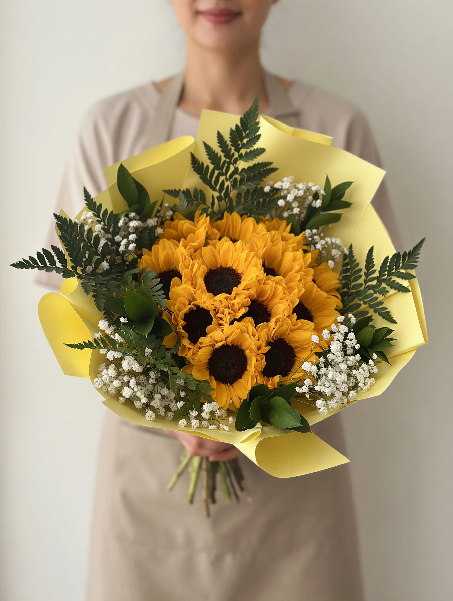 Sunflowers Bouquet Greenery / Ramo de Girasoles (10) - Bright, cheerful sunflowers take center stage in this sunflower bouquet - a lively everyday gift that feels like a pocket of summer. Perfect as a birthday bouquet, a thank-you arrangement, or a spring celebration surprise, the yellow sunflowers with their dark centers instantly make the person you're thinking of feel noticed and uplifted.  This hand-tied arrangement includes ten radiant sunflowers surrounded by small white filler blooms and rich green foliage for depth and contrast. The stems are finished in a pale yellow paper wrap that gives the bouquet a fresh, casual look and makes it easy to carry or present. The mix of bold yellow, crisp white, and leafy green creates a sunny, textured display that brightens any room.  Choose this bouquet to celebrate a friend, say thanks, or simply send warm, sunny wishes - it's the kind of gift that reads as joyful and sincere. For best enjoyment, trim stems and change the water every couple of days; the compact, rounded design makes it ideal for medium vases and easy to place around the home.