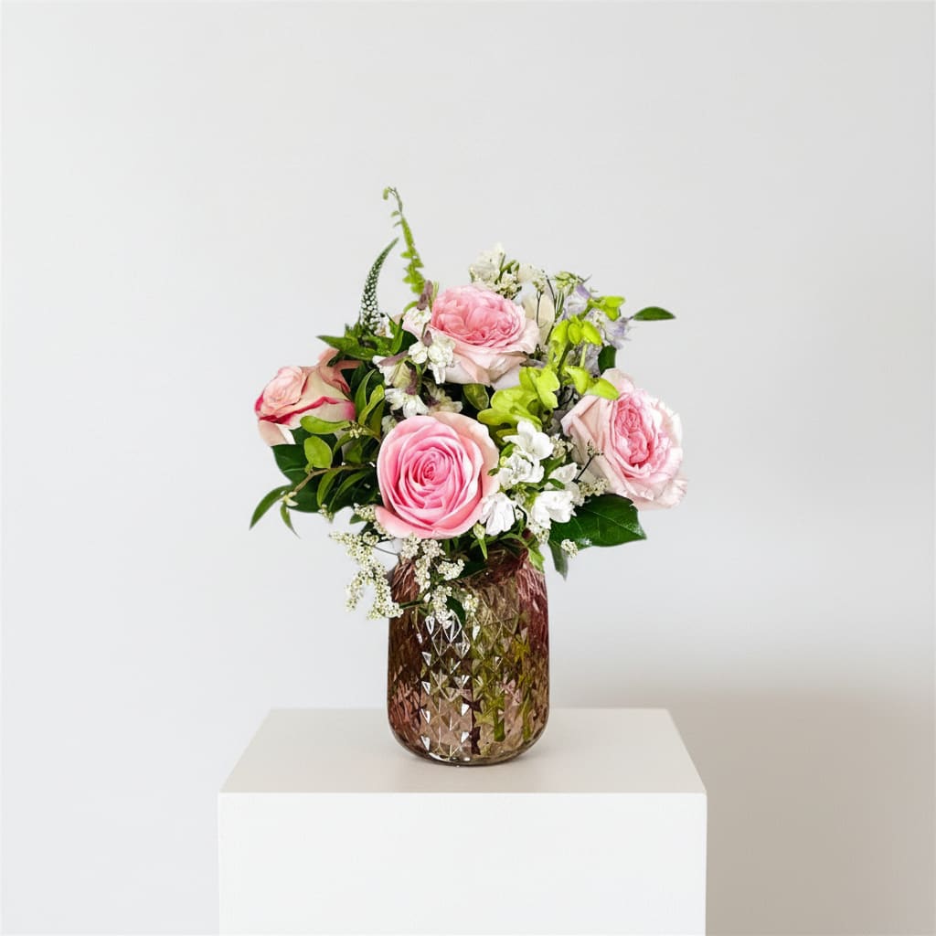 Whispers of the heart/ Luxurious Pink Roses Bouquet  - Soft pink roses gathered with delicate white stock and pale green orchids create a whisper-soft bouquet perfect as a Mother's Day flower arrangement or a gentle everyday gift. The romantic mix of pink, white, and green reads feminine and uplifting-the kind of arrangement that makes the person you're thinking of feel cherished and seen.  The blooms are artfully arranged in a textured faceted pink glass vase that adds subtle sparkle and a touch of vintage charm to tabletops or bedside tables. Large roses provide a lush focal point, while clusters of white stock and light-green orchid accents bring airy texture and soft contrast. Sized to brighten a small room, this design arrives ready to display; change water and recut stems every few days to extend freshness.