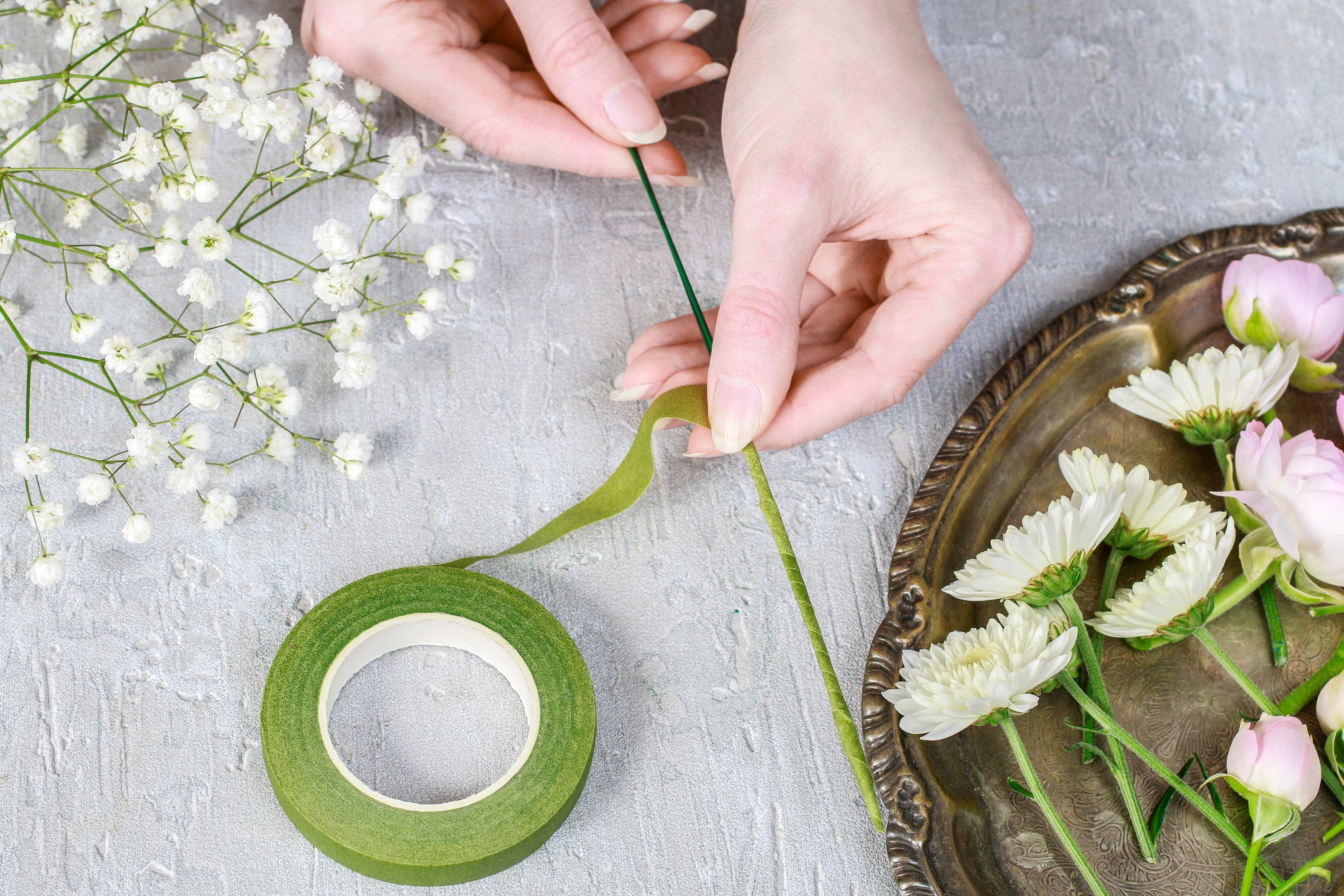 Hands wrapping flower stems with green floral tape beside small white and pink flowers