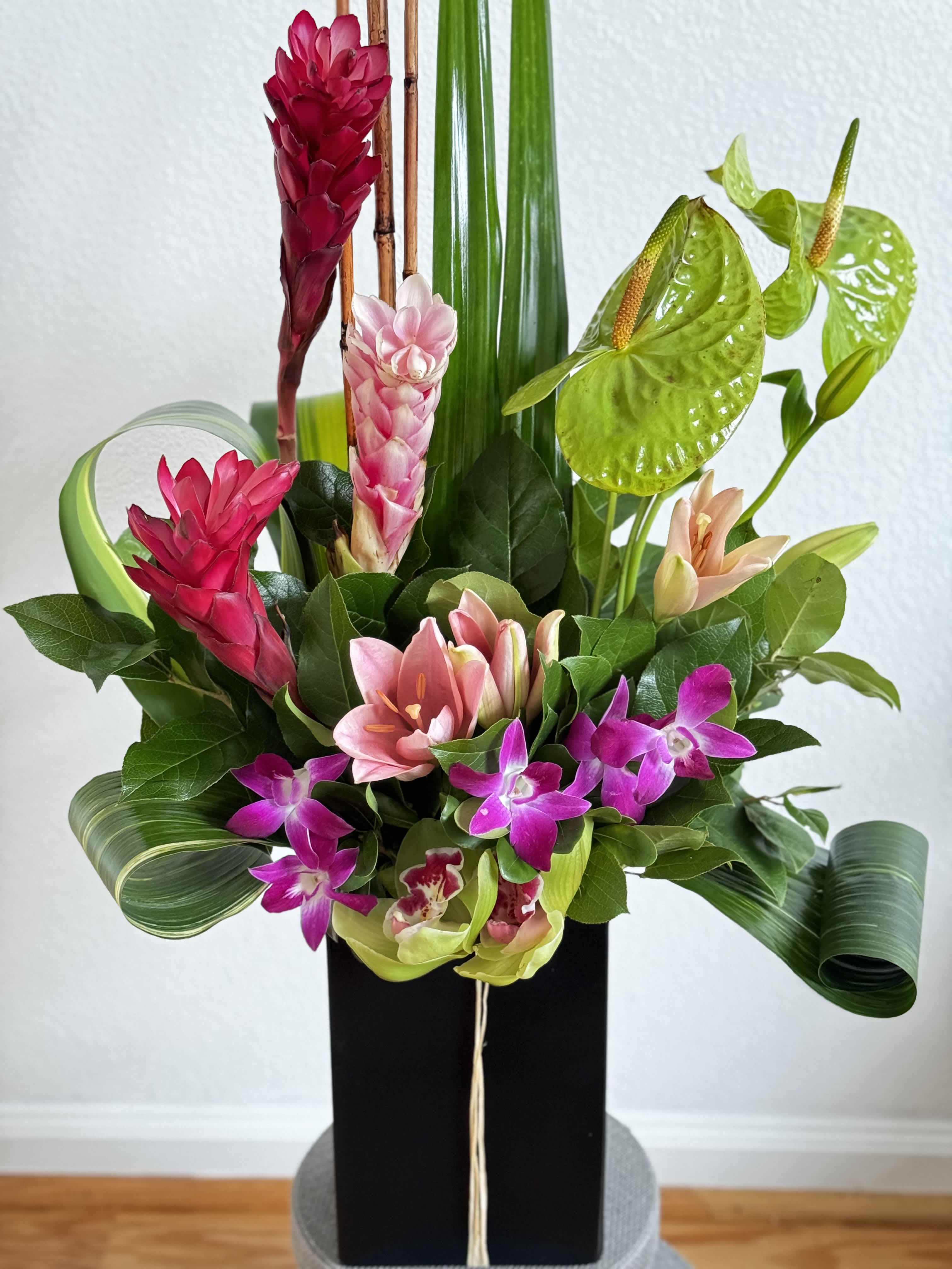 Tall tropical arrangement with pink lilies, orchids, ginger flowers, and sculpted green leaves in a black vase