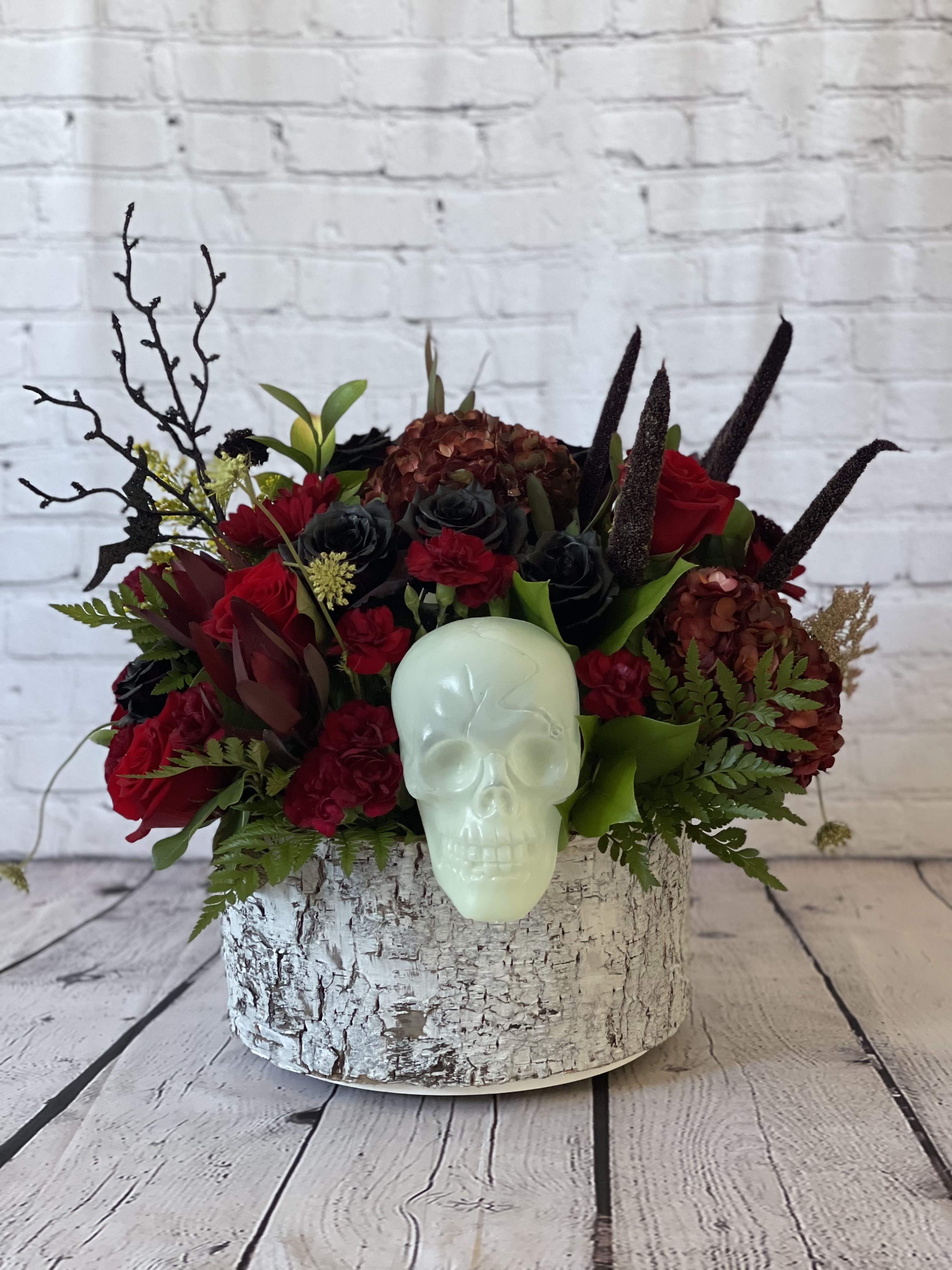 Gothic Garden  - Step into the dark beauty of the Gothic Garden with this haunting floral creation. Featuring a striking contrast of red and black roses intertwined with vibrant red hydrangea blooms, this arrangement exudes a mysterious allure. A white skull nestled among the lush blooms adds a macabre touch, creating an elegant yet eerie aesthetic. Housed in a rustic wood bark container, this Gothic Garden arrangement stands as a dramatic centerpiece, perfect for adding a touch of gothic romance to any space.