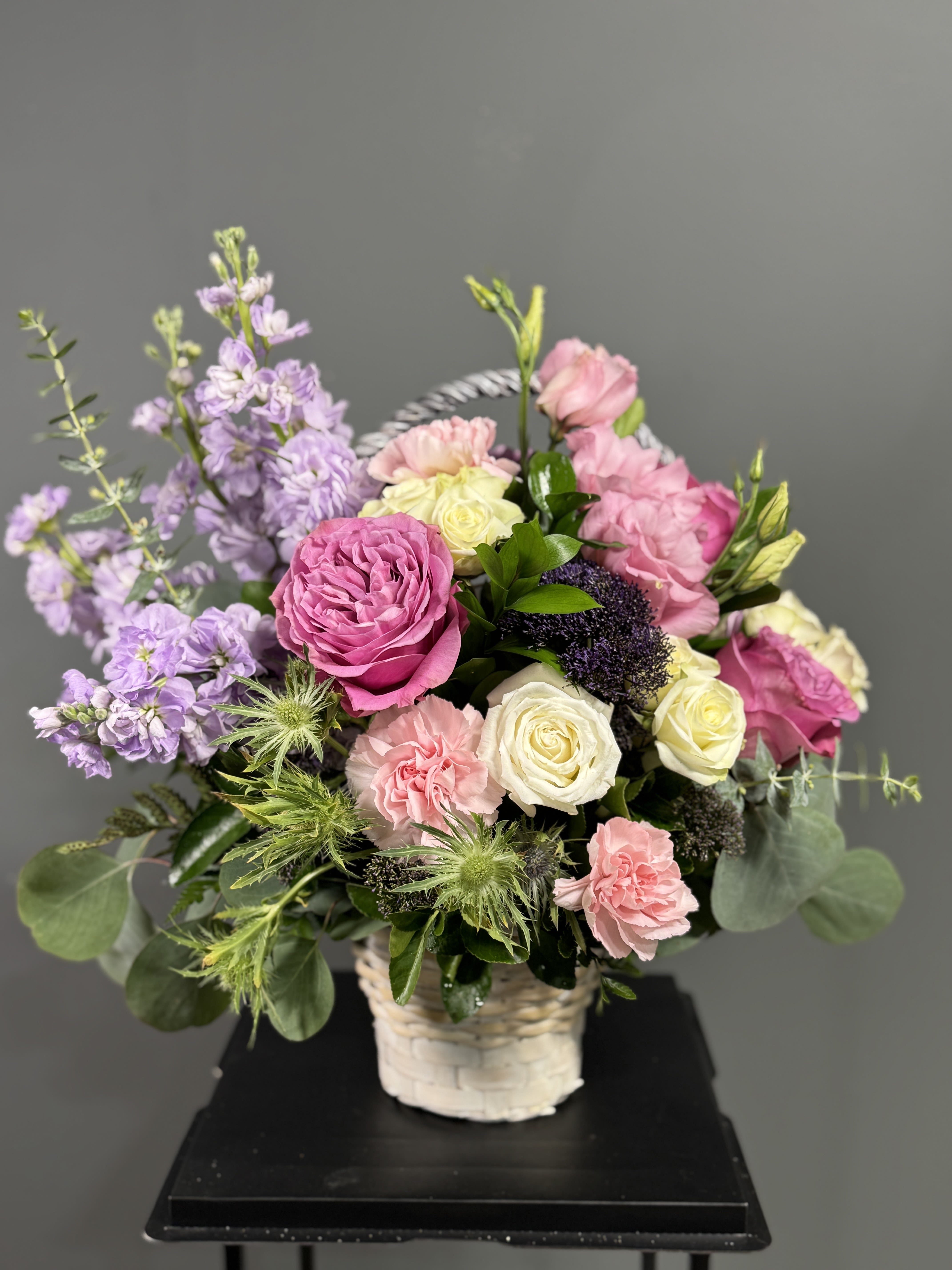 Basket arrangement of pink and cream roses with lavender stock and green thistle blooms