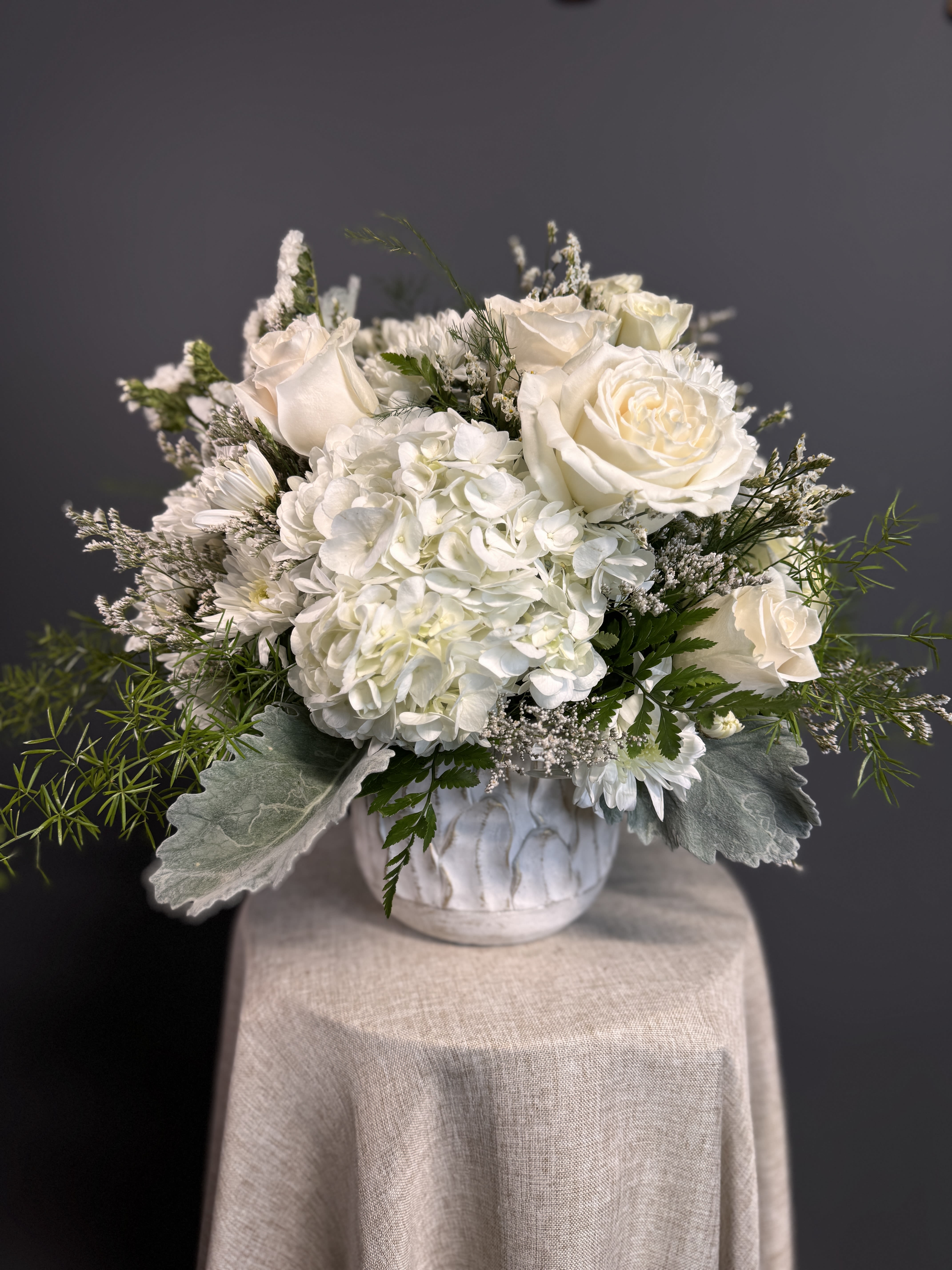Low white arrangement of roses and hydrangeas in a white ceramic pot on a draped pedestal