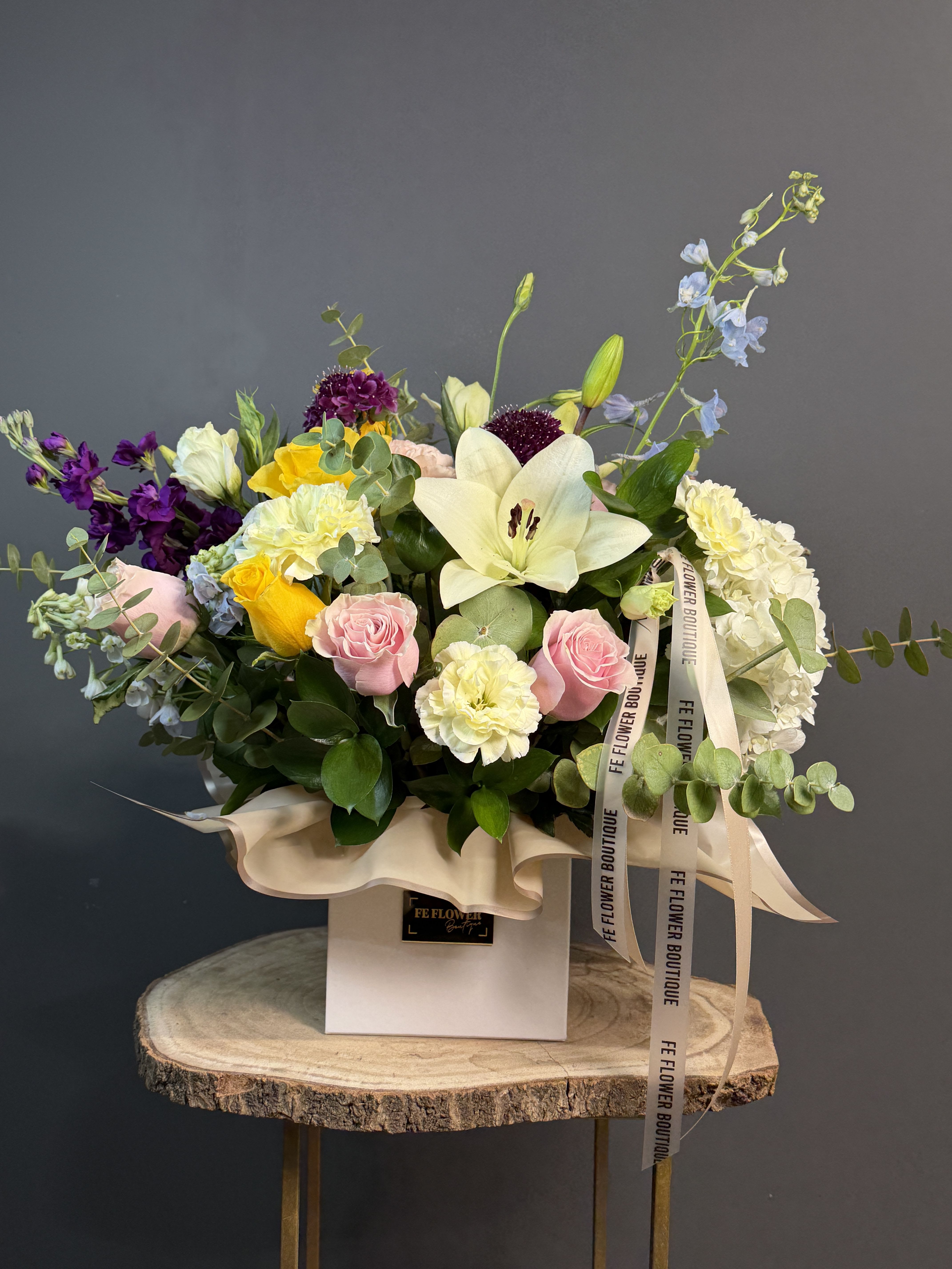 Colorful mixed flower arrangement with lilies, roses, and carnations in a white box on a wood stand.
