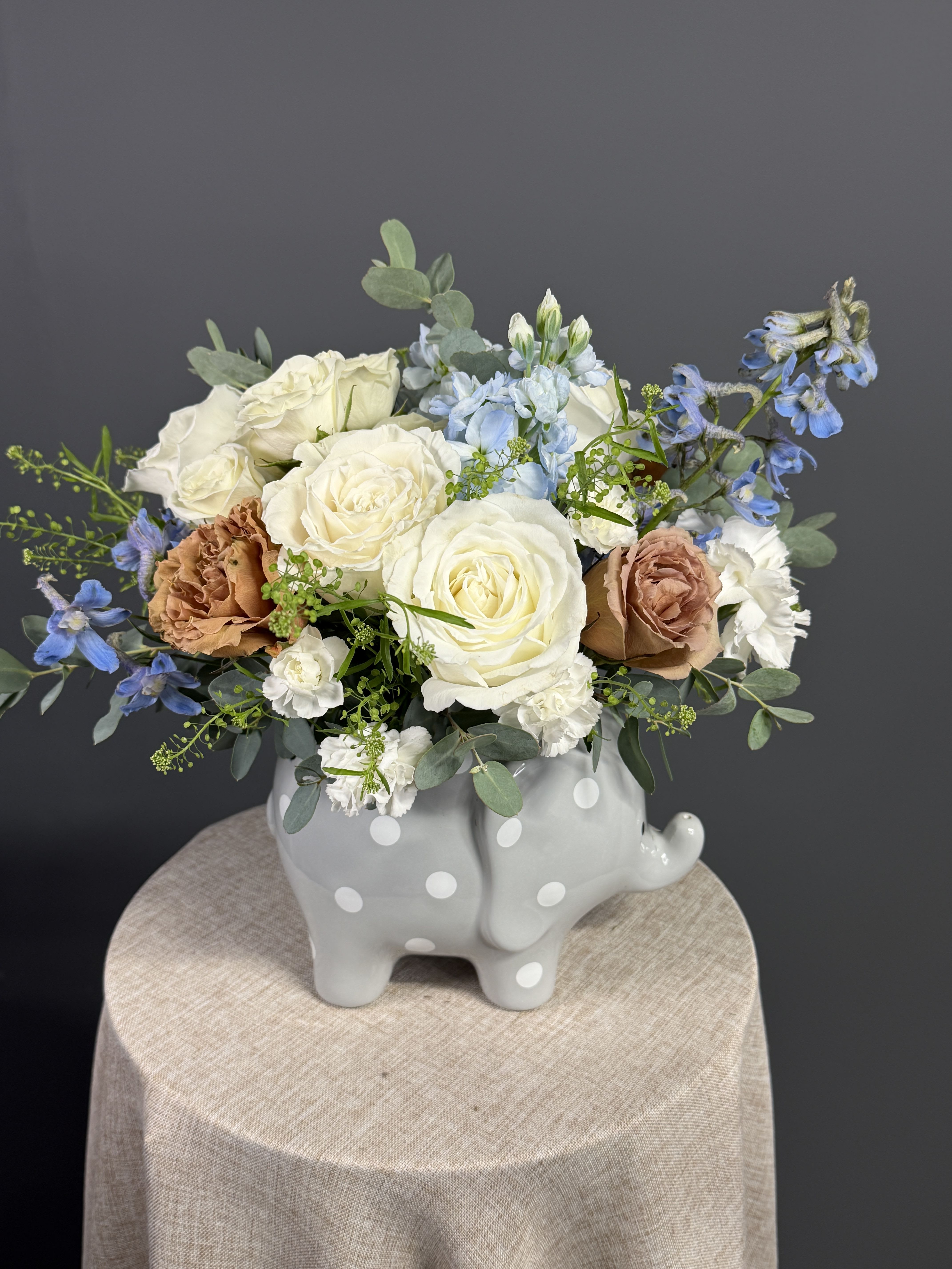 Blue Baby Snuggles Elephant - A charming baby boy arrangement featuring soft blue blooms, creamy white roses, and delicate textures, beautifully designed in a keepsake elephant vase. Blue Baby Snuggles Elephant is a sweet and thoughtful way to welcome a little one with love and gentle elegance