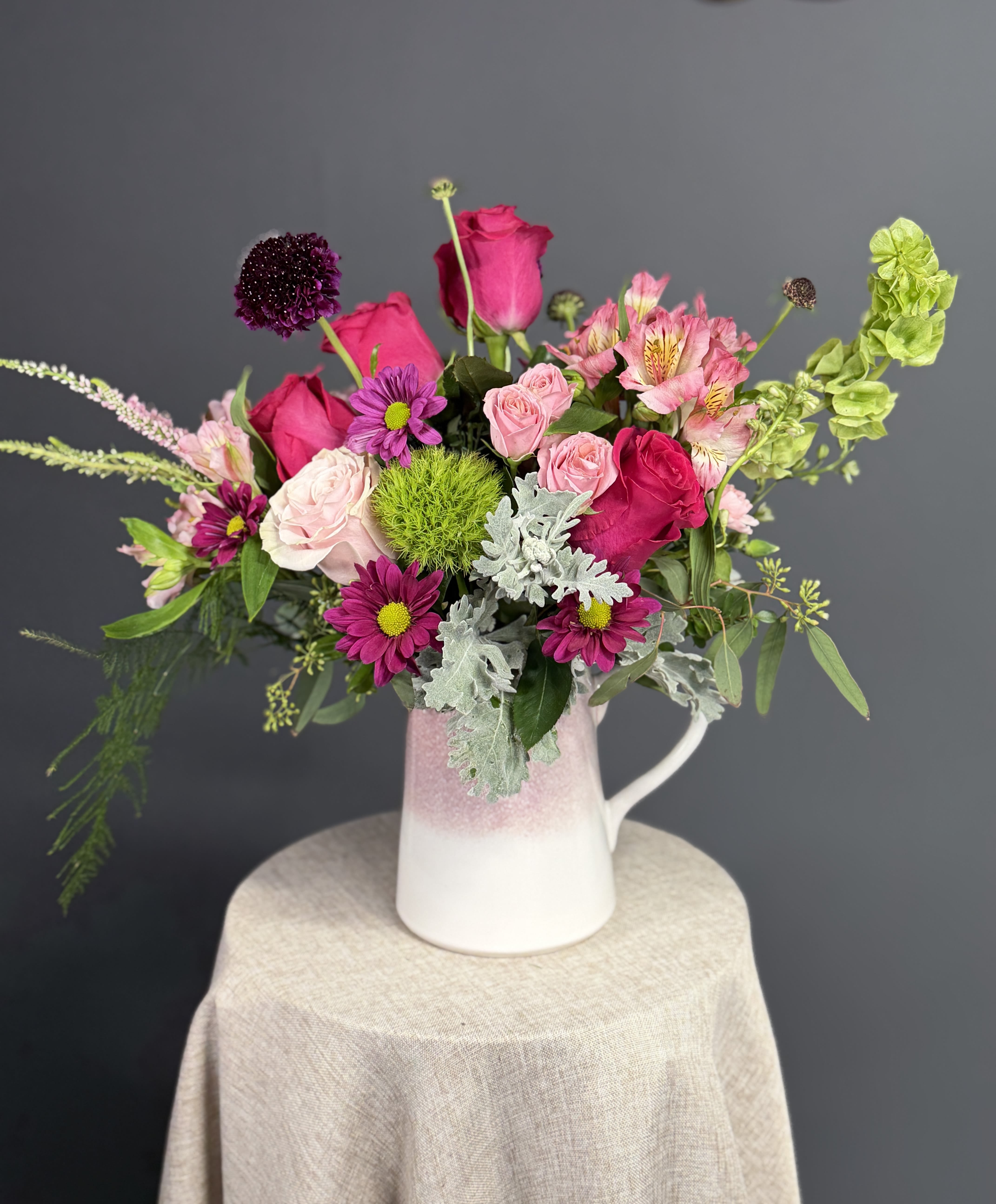 Berry Sorbet Soirée - Capturing the untamed beauty of a wild meadow, this vibrant arrangement is a celebration of texture and color. Anchored by luxurious deep magenta roses and delicate blush spray roses, the design is accented with striking dark purple scabiosa and a centerpiece of neon green dianthus. Softening the bold jewel tones are whimsical sprigs of pink heather, silvery dusty miller, and a variety of eucalyptus and fern foliage that add a natural, airy movement. Presented in a charming, two-tone stoneware pitcher with a soft mauve ombre finish, this bouquet is as much a keepsake as it is a floral statement.