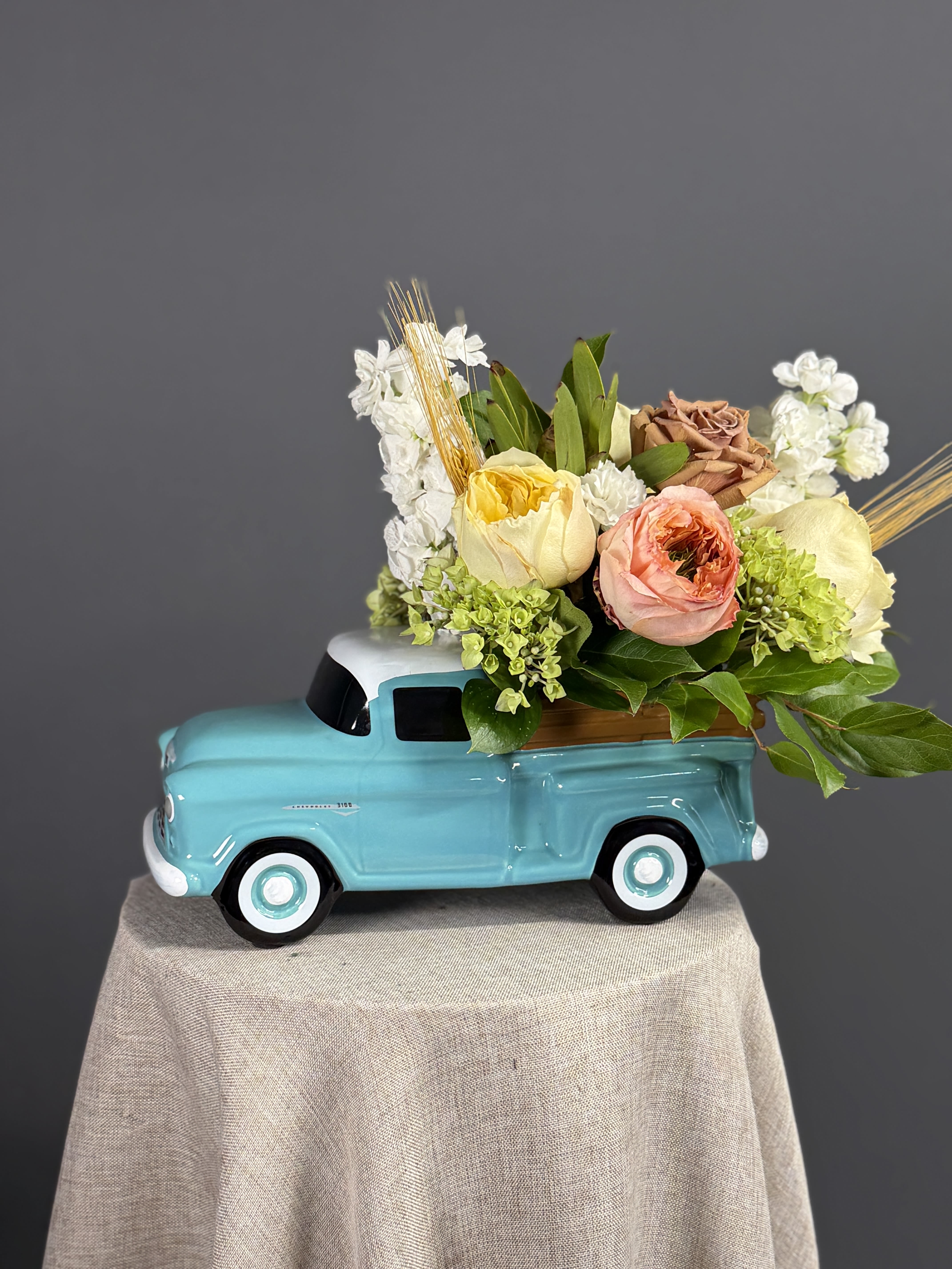 The Teal Hauler - A glossy tale ceramic Ford pickup holds a cheerful, tightly packed cluster of roses, hydrangea accents and blooms in soft coral, creamy whites, buttery yellows and warm tan tones, accented with broad green leaves and decorative dried wheat for a whimsical, slightly rustic feel-an everyday flower arrangement perfect for birthdays, housewarmings, thank-you gifts or a lighthearted thinking-of-you surprise that also doubles as a charming keepsake.   If substitutions are needed, we'll match colors and textures to preserve the same playful vintage look.