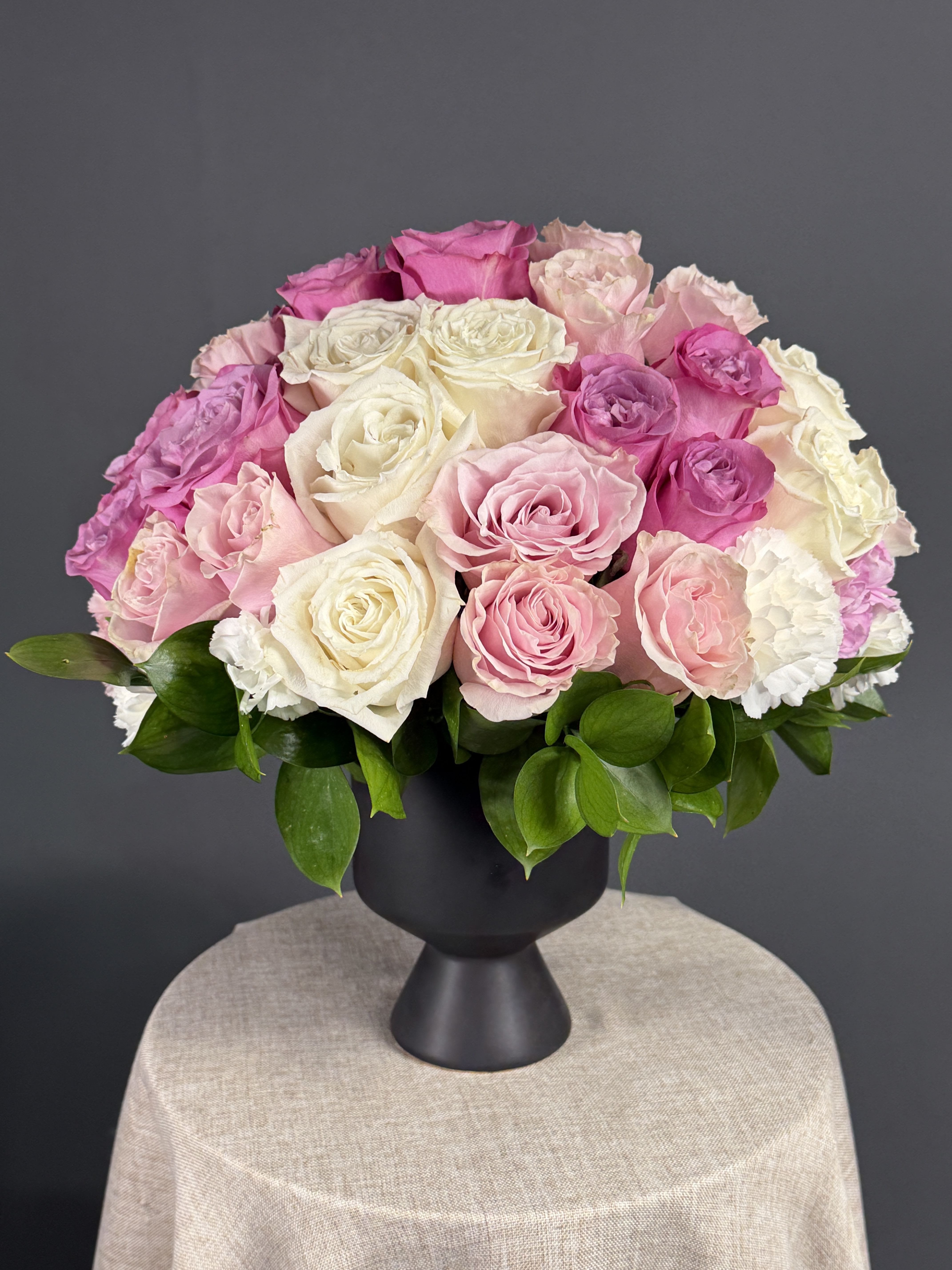 Whispers of Love  - A lush dome of blush pink, hot pink, creamy white and soft lavender roses sits against a matte black pedestal vase for a polished, romantic birthday bouquet or anniversary arrangement that makes the person you're thinking of feel genuinely celebrated. Tightly packed blooms form a full, rounded silhouette with glossy green foliage peeking at the base, the dark vase sharpening the pastel palette for Mother's Day, spring celebrations, or an elegant everyday gift.   If a specific rose color is unavailable, substitutions of equal quality and tone will be made to preserve the overall look and sentiment.