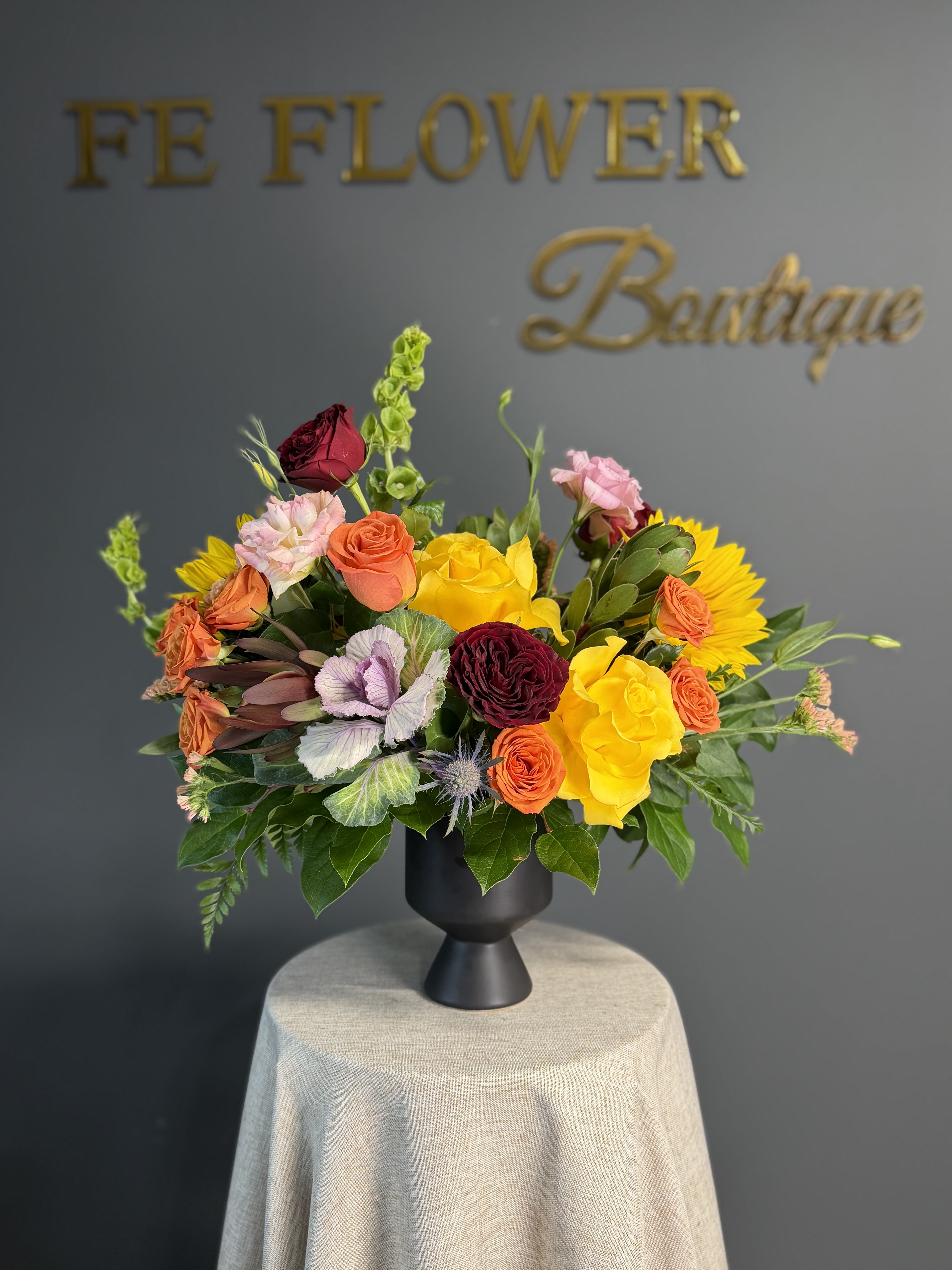 Bloom Drama - A vivid, sculptural arrangement built for celebration: Bloom Drama is a bold flower arrangement perfect for an anniversary, birthday, Mother's Day gift, or a cheerful just-because surprise. Sunflowers and orange-yellow roses meet deep burgundy roses and soft pink spray roses for a warm, upbeat palette that signals joy and care the moment someone sees it.  Set in a matte black ceramic pedestal vase, the composition adds height and movement with Bells of Ireland and variegated ornamental kale, while spiky blue thistle brings an earthy, contemporary edge. Sized to sit confidently on a round table or sideboard, it arrives ready to display on a fabric-covered surface and is an excellent pick for thank-you, get-well, thinking-of-you, Women's Day, or congrats moments. Substitutions may be made with flowers of equal value to preserve the overall style and color story.