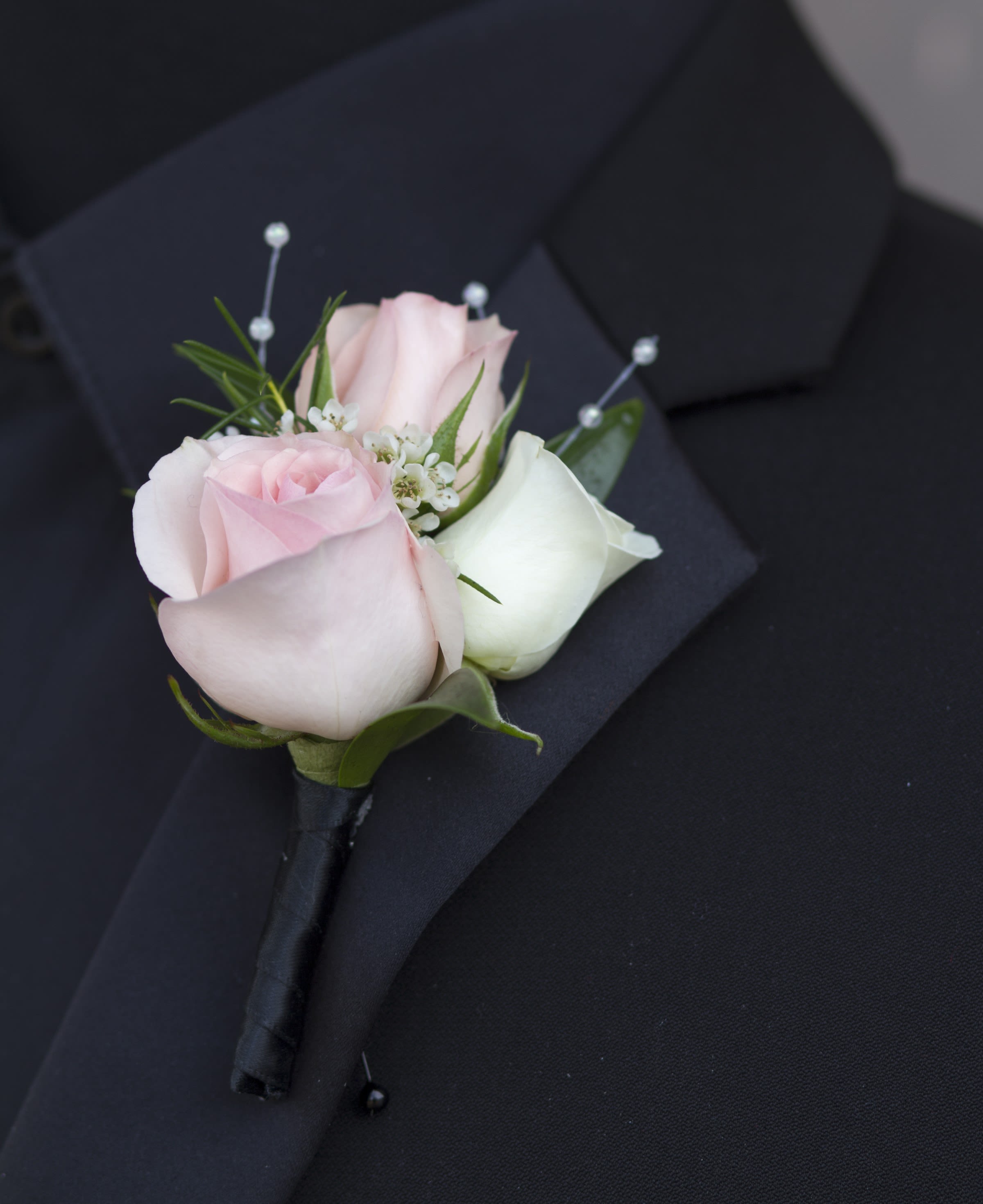 Asheville - A cluster of pale pink and white spray roses forms a compact boutonniere that reads romantic and refined - perfect as a prom corsage or for weddings and other dressy events. Small white filler blooms peek between the roses to add soft texture and a delicate contrast, while pearl accents catch the light for a subtle, formal sparkle that makes the recipient feel celebrated.  Stems are wrapped in a sleek black ribbon for a tailored finish, with the pearl pins securing the design and enhancing the classic look. If you prefer a different stem wrap color, include that request in the special instructions at checkout.  Pick up dates for prom are Saturday, April 11th, 18th, 25th and May 2nd; please call if you need this arranged for a different event.