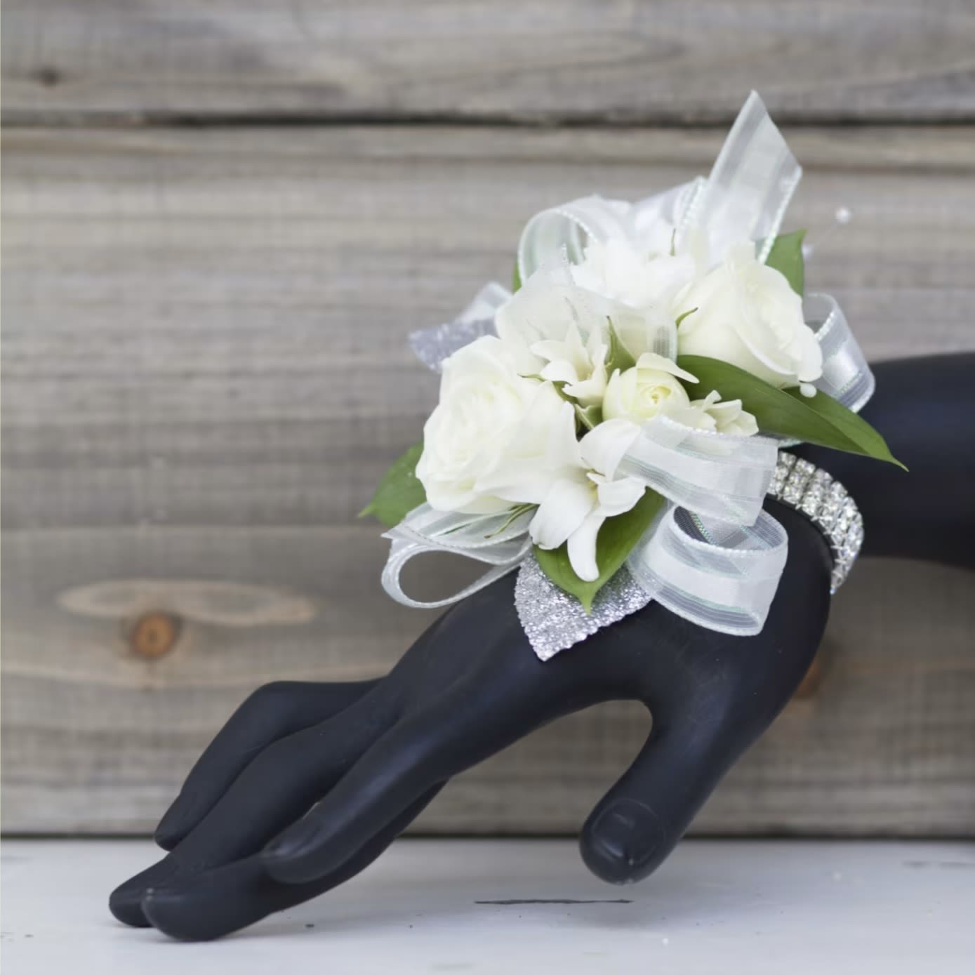 Athens - Classic, couture white blooms, hand selected by our professional designers; arranged with shimmering silver accents make this prom corsage a striking choice for formal occasions. The wrist corsage reads like a piece of jewelry-clusters of white focal blossoms and petite accent blooms sit against loops of sheer ribbon and a silver accents on a rhinestone bracelet band-so the person you're honoring will feel both polished and noticed. Upgrade to deluxe and include a matching boutonniere~ a compact cluster of white roses and a coordinating white bloom, finished with a black, ribbon-wrapped stem for a tailored, tuxedo-ready contrast.  Carefully constructed for comfort and sparkle, the corsage rests on a flexible rhinestone or pearl bracelet that slips on easily and shows beautifully against dark or light formal wear; the boutonniere pins securely to a lapel for a crisp, photo-ready look. This pairing suits prom, weddings, quinceañeras, anniversaries, or any special celebration when you want her to feel celebrated and elegant.   Pickup dates for prom are Saturday, April 11th, 18th, 25th, and May 2nd-please call if you need these for a different event.