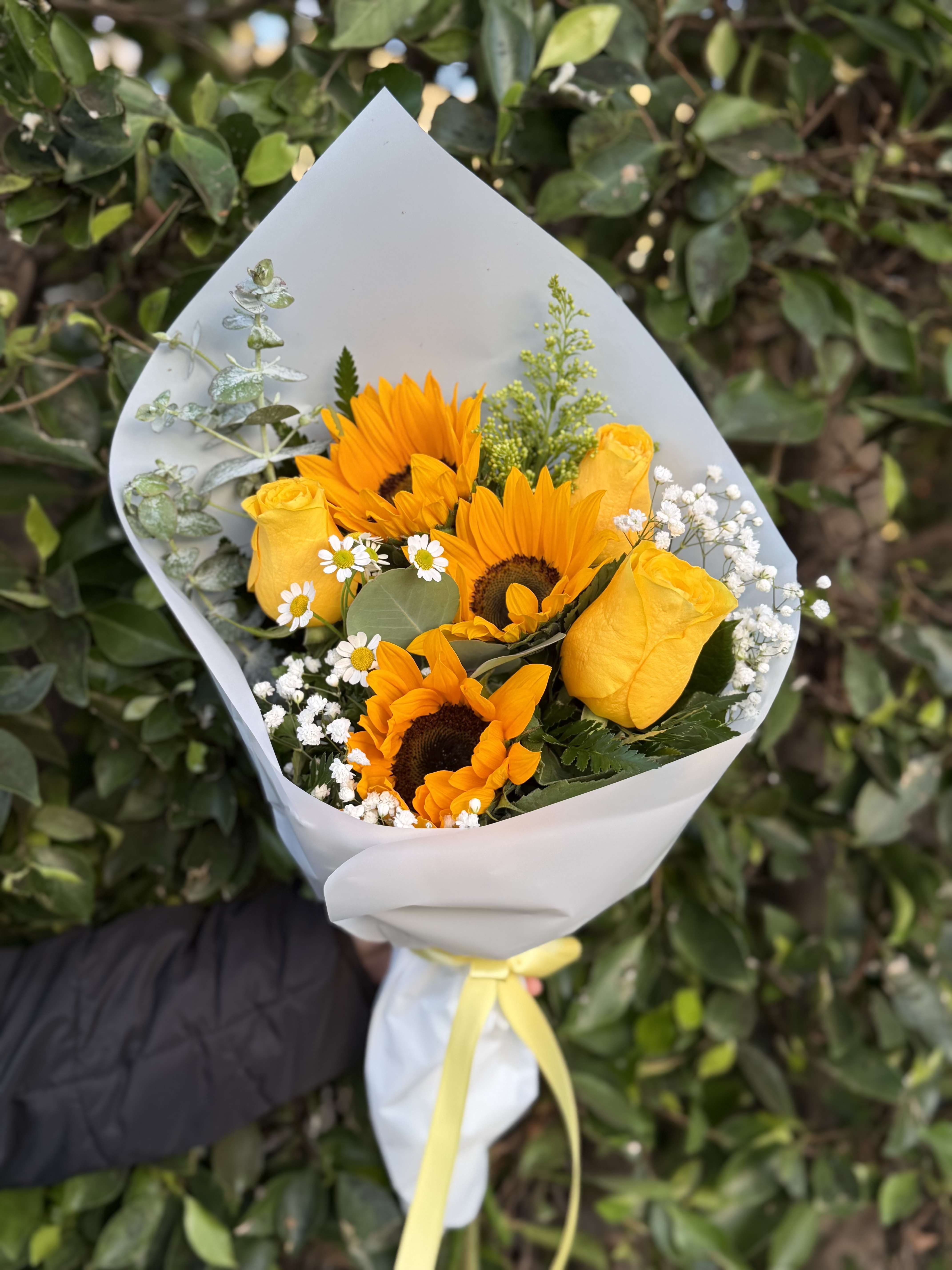 Hand-tied bouquet of bright yellow sunflowers and roses wrapped in white paper with a yellow ribbon