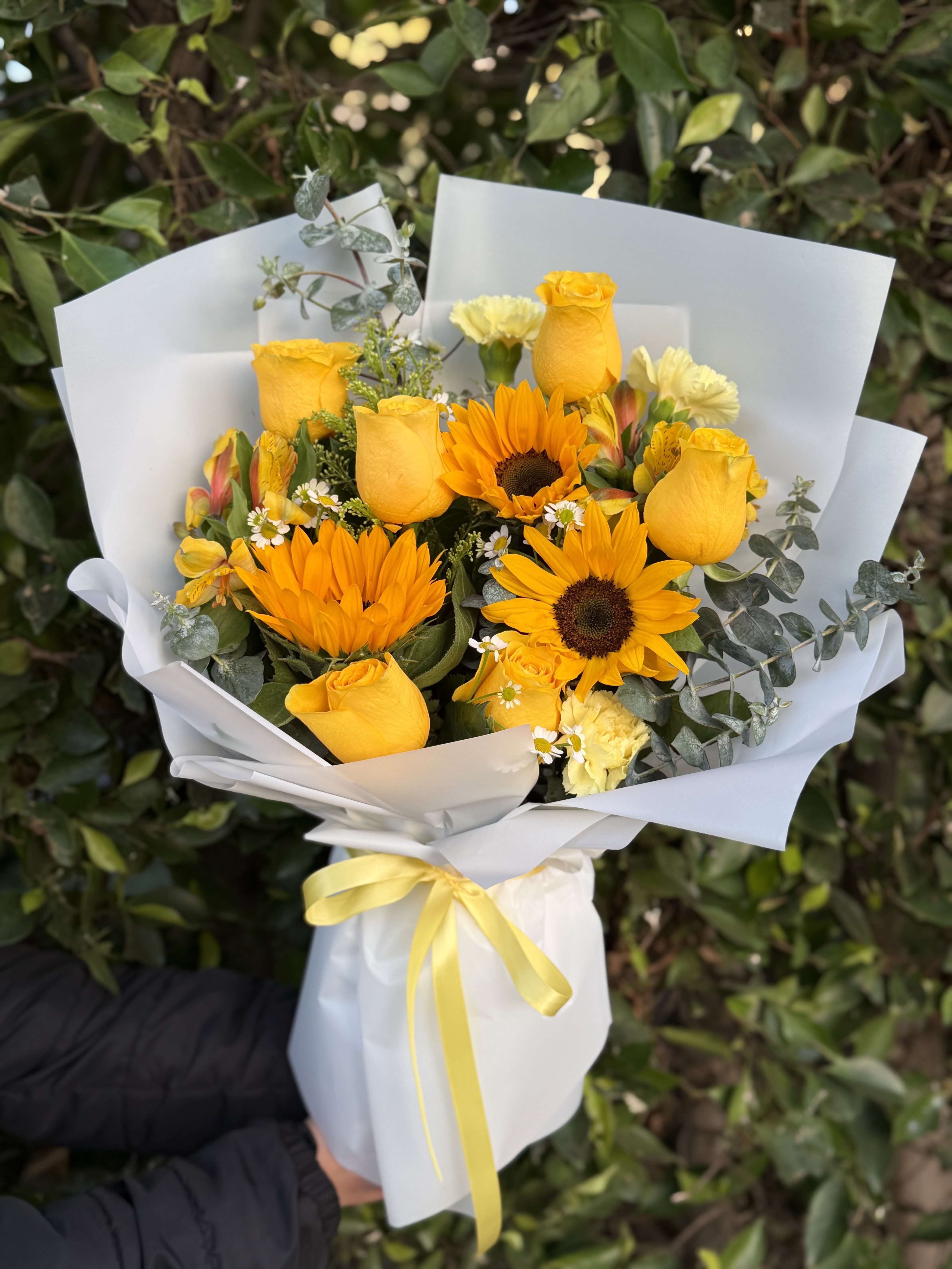 Sunlit Radiance  - A luminous one-faced luxury wrapped bouquet of sunflowers and golden yellow roses, thoughtfully arranged with delicate filler blooms and fresh eucalyptus. Designed in a front-facing, layered style and finished in an elevated wrap with a soft satin ribbon, Sunlit Radiance is presented with a coordinating glass vase—offering a bold yet refined presentation that is warm, glowing, and effortlessly elegant.