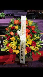 Standing funeral spray of red, yellow, and orange flowers with a white ribbon