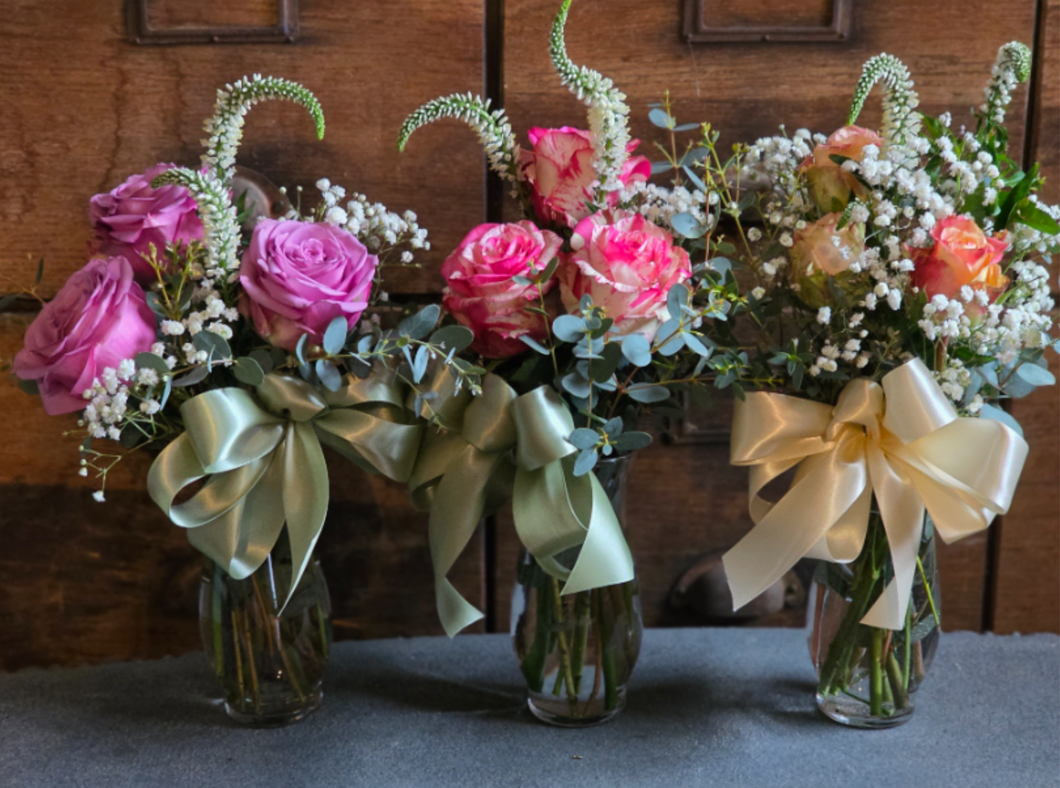 Three small glass vases with pink and peach roses, white fillers, and large satin ribbon bows.