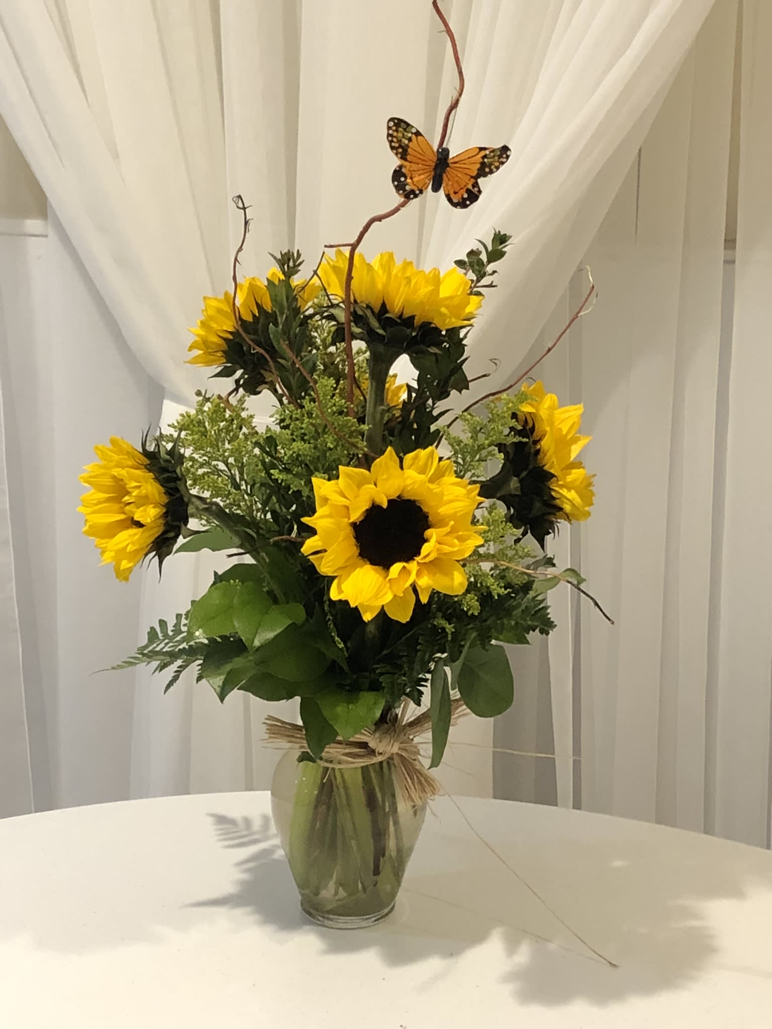 Medium vase of bright yellow sunflowers with accent fillers and a decorative butterfly on curly branches