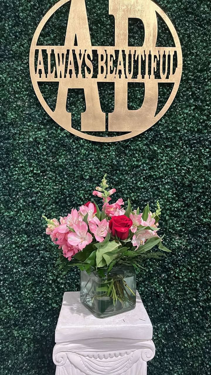 Pink flowers and a red rose in a glass vase beneath a gold "Always Beautiful" sign