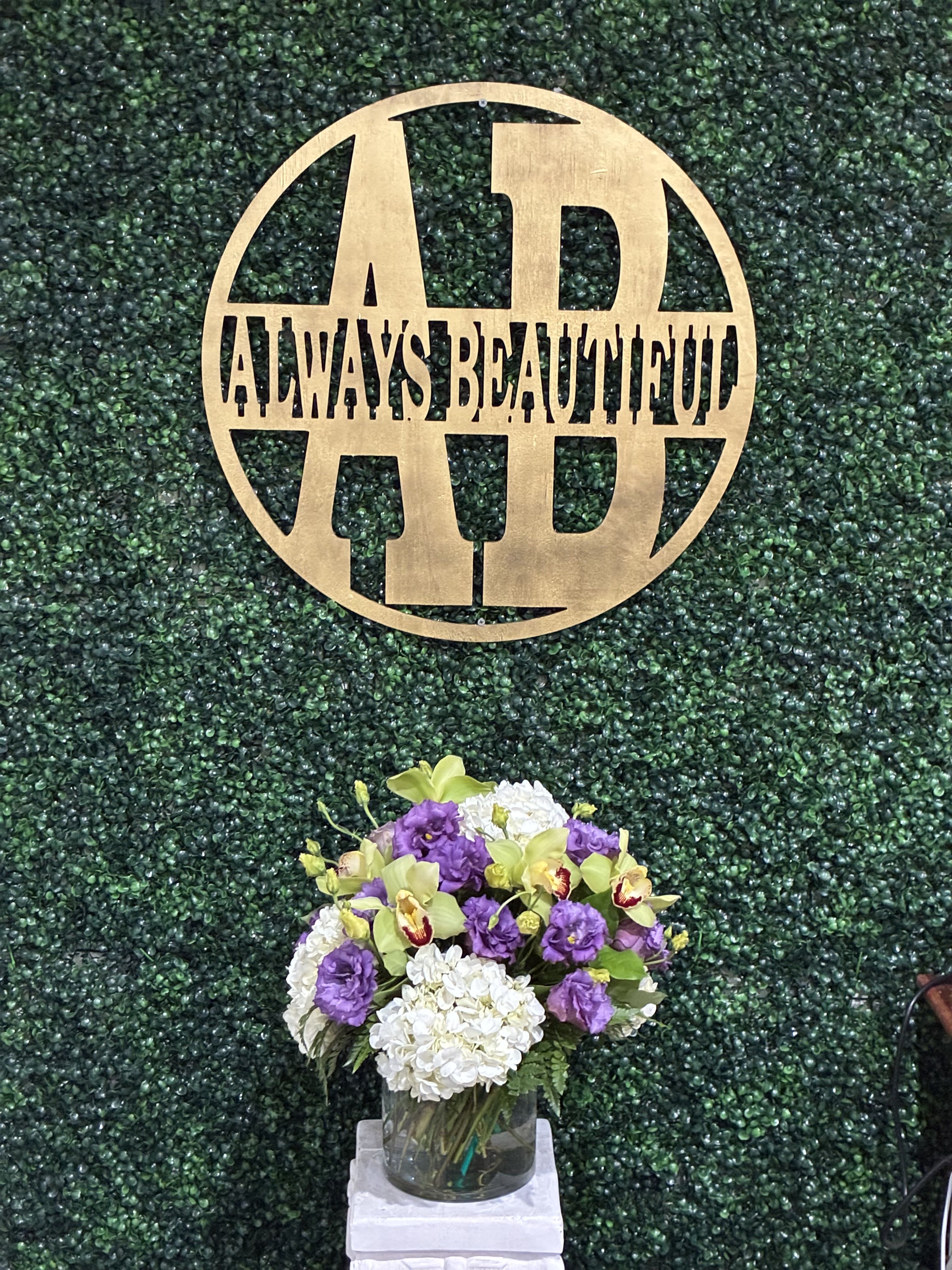 Purple and white floral arrangement in a glass vase beneath a gold monogram sign