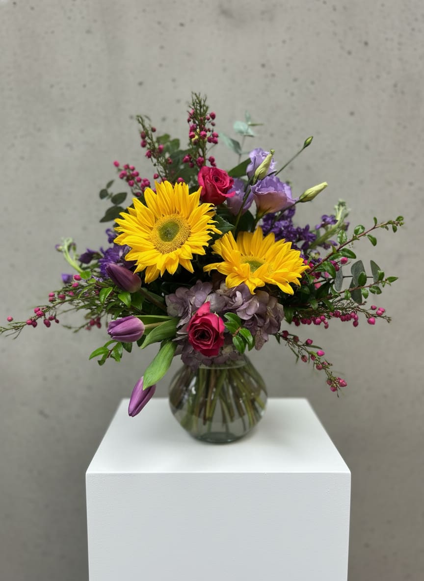 Good Day Sunshine  - Two bold yellow gerbera daisies take center stage in this mixed-color flower arrangement, creating an instant mood lift for a birthday, get well wish, housewarming, or just-because surprise. The bouquet reads bright and cheerful-perfect for spring and summer celebrations or to bring warmth into an autumn or Thanksgiving table-yet it still fits seasonal moments like Halloween when paired with warmer décor. The person you're sending it to will feel noticed and genuinely cheered by the vivid focal blooms and playful color mix.  Surrounding the gerberas are hot pink roses and lavender lisianthus that add romantic and soft-textured layers, while deep purple tulips and muted mauve hydrangeas give the design depth and contrast. Small pink berry-like accents and delicate filler flowers create a loose, airy silhouette that feels informal and lively. Arranged in a round clear glass vase that displays the stems fanning naturally, this bouquet is sized to sit comfortably on a bedside table or dining centerpiece.  This arrangement suits many occasions-anniversary, graduation, thank you, congratulations, sympathy or thinking-of-you moments-and also makes a bright new-baby or retirement gift. Care is simple: keep the vase filled with fresh water, trim stems every few days, and place away from direct heat or drafts to extend bloom life. Order with same-week delivery when available to make sure your recipient enjoys the freshest, sunniest welcome.