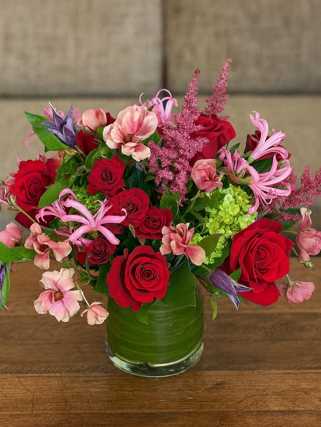 Velvety &amp; Crimson Harmony - A romantic medley of reds and crimson designed to make hearts flutter. This lush arrangement features velvety red roses, delicate blush butterfly ranunculus, and playful pink nerines accented with soft textures of astilbe and fresh greenery. Each stem is artfully composed in a clear glass vase wrapped with a green leaf lining — a modern touch that highlights the natural beauty within.  Perfect for anniversaries, special celebrations, or simply to say “I’m thinking of you,” this bouquet captures passion and elegance in one vibrant statement.   Available exclusively at Sunny Florists – handcrafted with love, designed to delight.