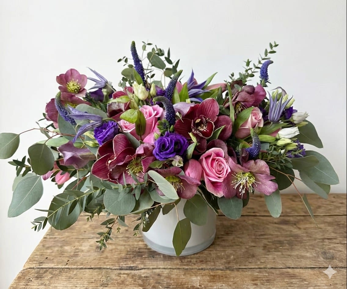 Burgundy Spring - This arrangement is present in a 6"*6" white cylinder vase. The arrangement includes burgundy color orchid, hellebores, purple color veronica, clematics, lisanthus, calla lily and pink tulip and roses. 
