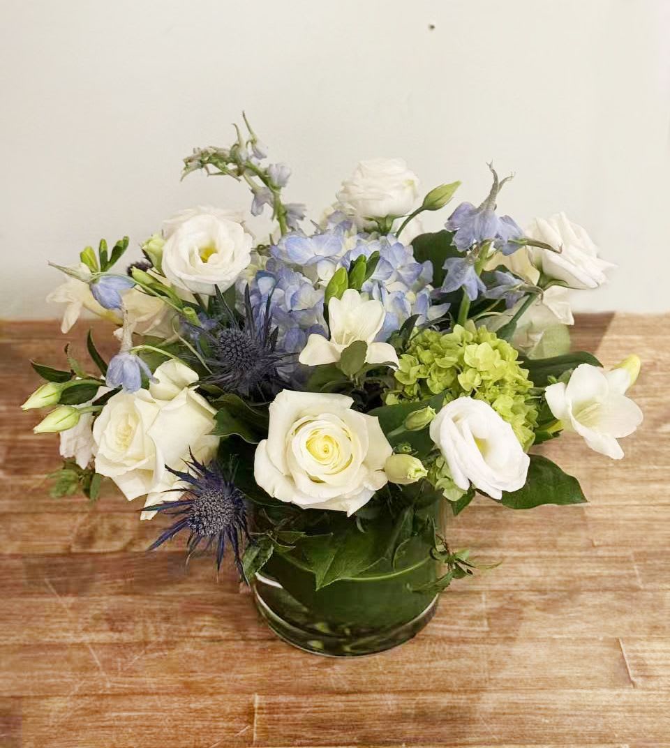 Annie - Elegant and refreshing, this soft blue-and-white arrangement brings a calm, garden-inspired feel to any space. Creamy white roses and lisianthus are artfully paired with airy blue delphinium, delicate hydrangea, and sculptural blue thistle, all accented with lush seasonal greenery. Designed low and full in a clear glass cylinder vase, this piece feels effortless yet refined.  Perfect for birthdays, thank-you gifts, sympathy gestures, or simply brightening a home or office, this arrangement offers a serene palette and natural movement that feels both modern and timeless.  Details: 	•	Features white roses, white lisianthus, blue delphinium, blue thistle, and hydrangea 	•	Arranged in a clear glass cylinder vase 	•	Soft, airy design with a fresh garden look 	•	Designed by Sunny Florists  Please note: Flower varieties may vary slightly due to seasonal availability, while maintaining the overall color palette and design style.
