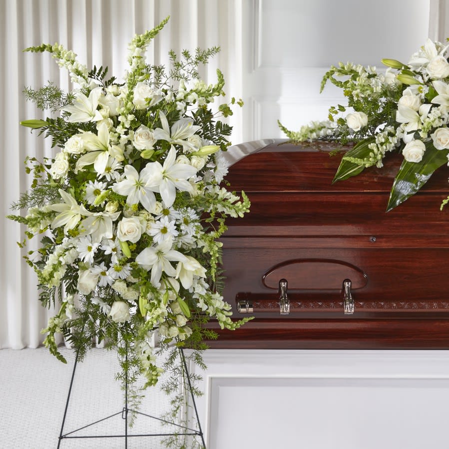 Tall white funeral spray with lilies, roses, and daisies on a stand beside a wooden casket.