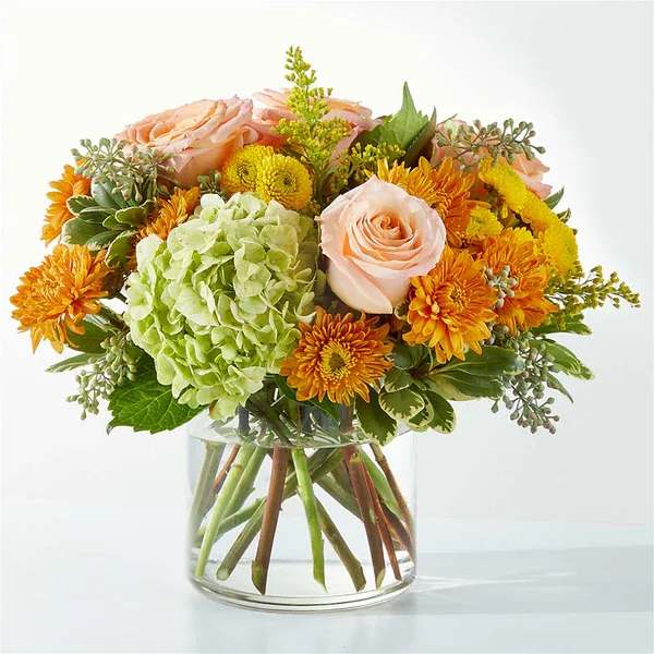 Round bouquet of peach roses, orange mums, and green hydrangea in a clear glass cylinder vase