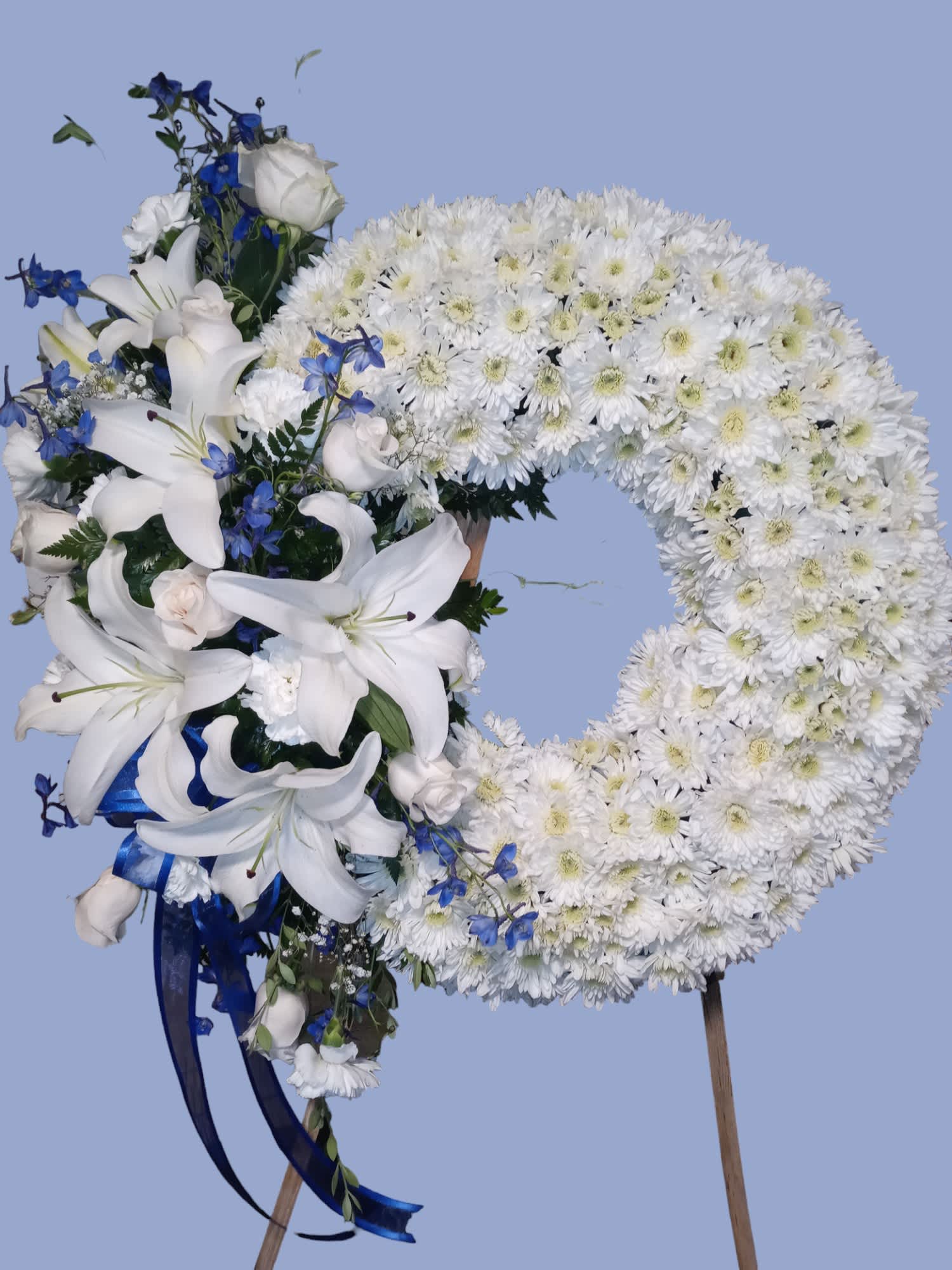White floral wreath with lilies and blue accents on a stand