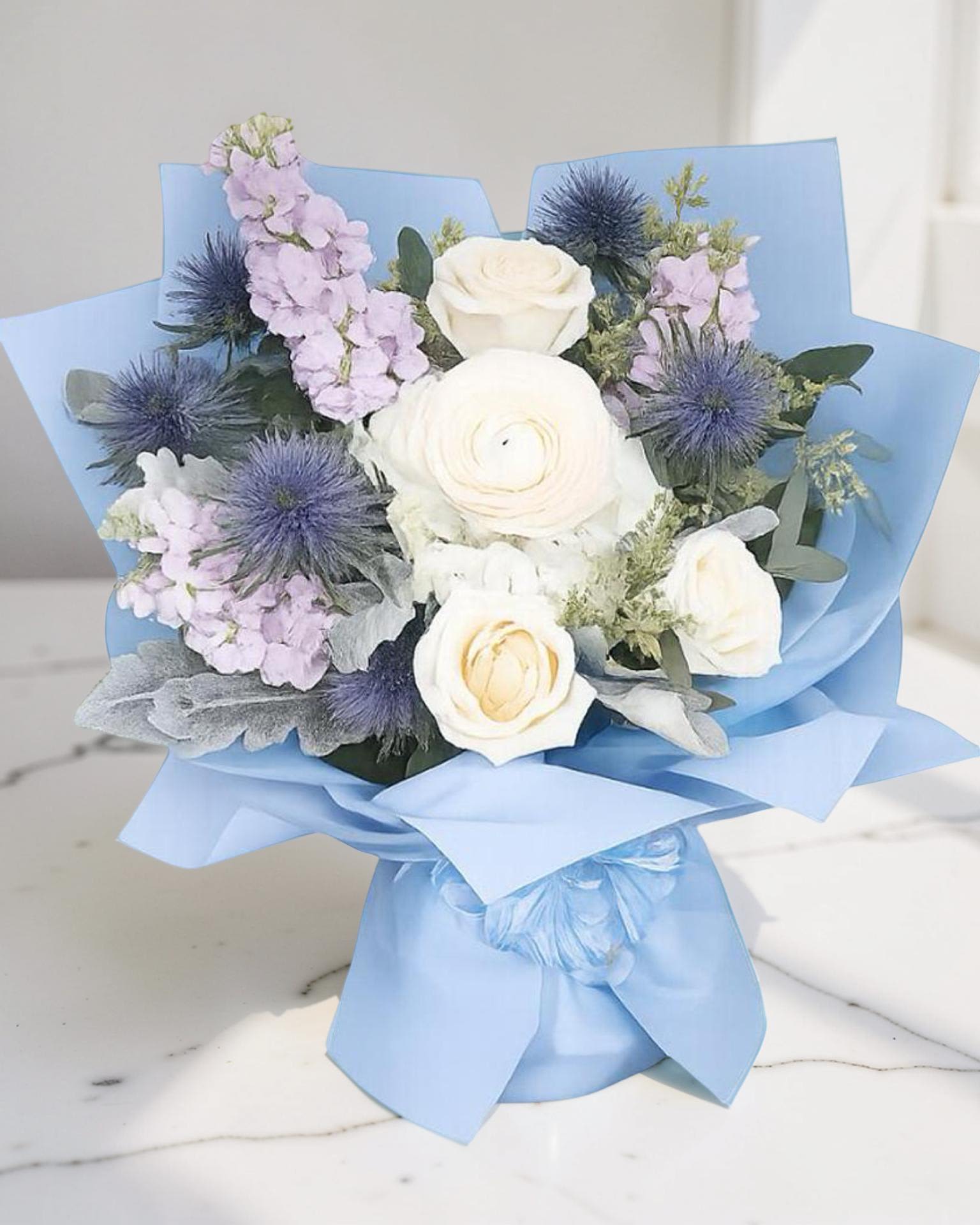 Columbia Blue Celebration Bouquet - A beautiful hand-wrapped inspired by Columbia’s signature blue, featuring elegant white roses, soft lavender blooms, and striking blue thistle, wrapped in matching blue tones. Designed to celebrate achievement and new beginnings, this bouquet is perfect for Columbia University graduations. A fresh, modern design that feels polished, meaningful, and far from ordinary—just like the milestone it honors.