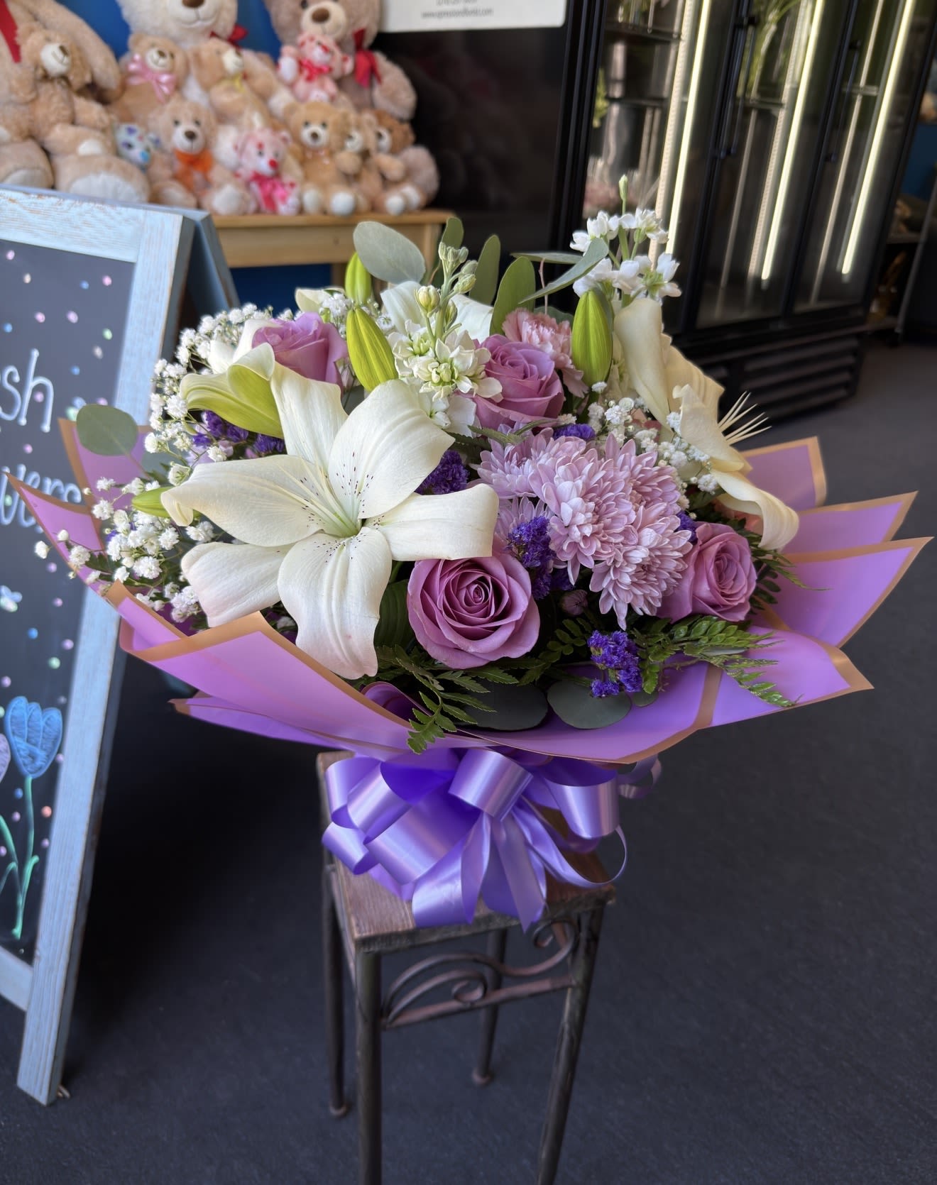 Purple Xpression - White lilies and lavender roses with pink chrysanthemums and greenery, wrapped in lilac paper and purple ribbon - a soft, romantic bouquet for birthdays, anniversaries, and spring celebrations.