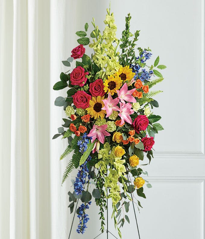 Love Lives on spray - Tall cascading spray of hot pink roses, yellow sunflowers, pink lilies and blue accents on a standing easel-vibrant, heartfelt tribute for memorials and remembrance.
