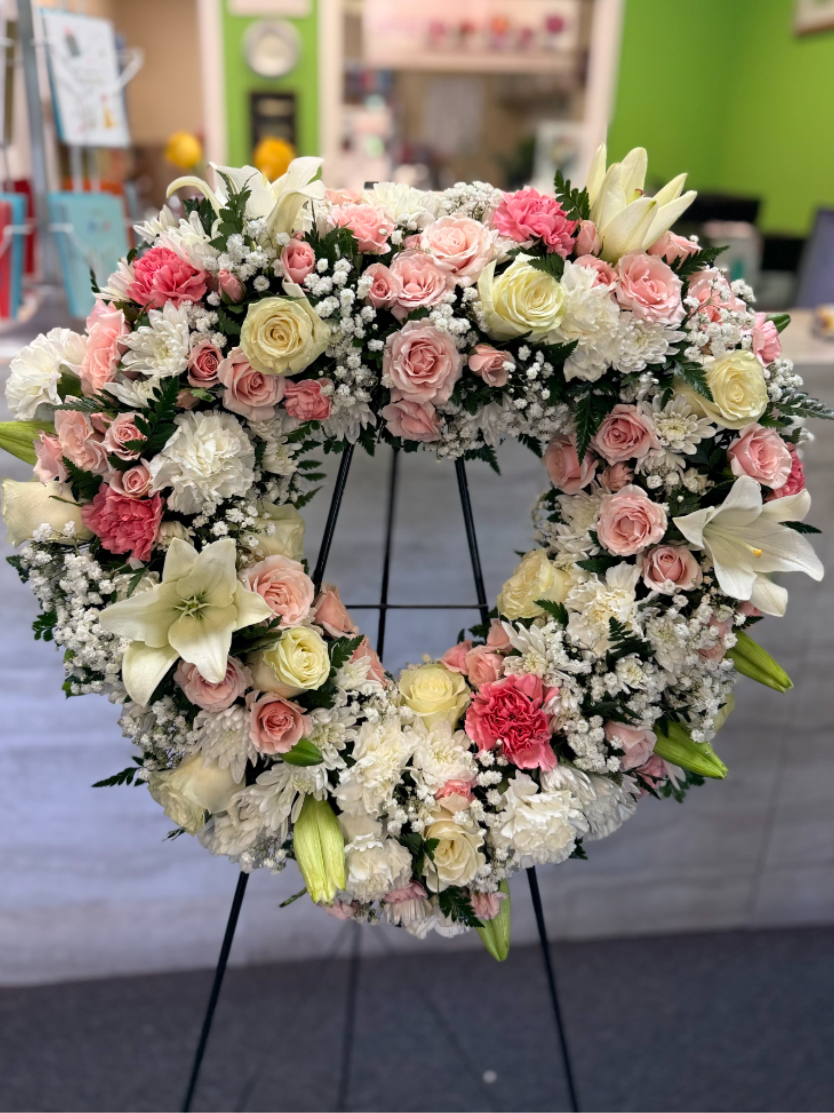Heart-shaped standing wreath of white, pink, and cream flowers with roses, lilies, and carnations on a black easel.