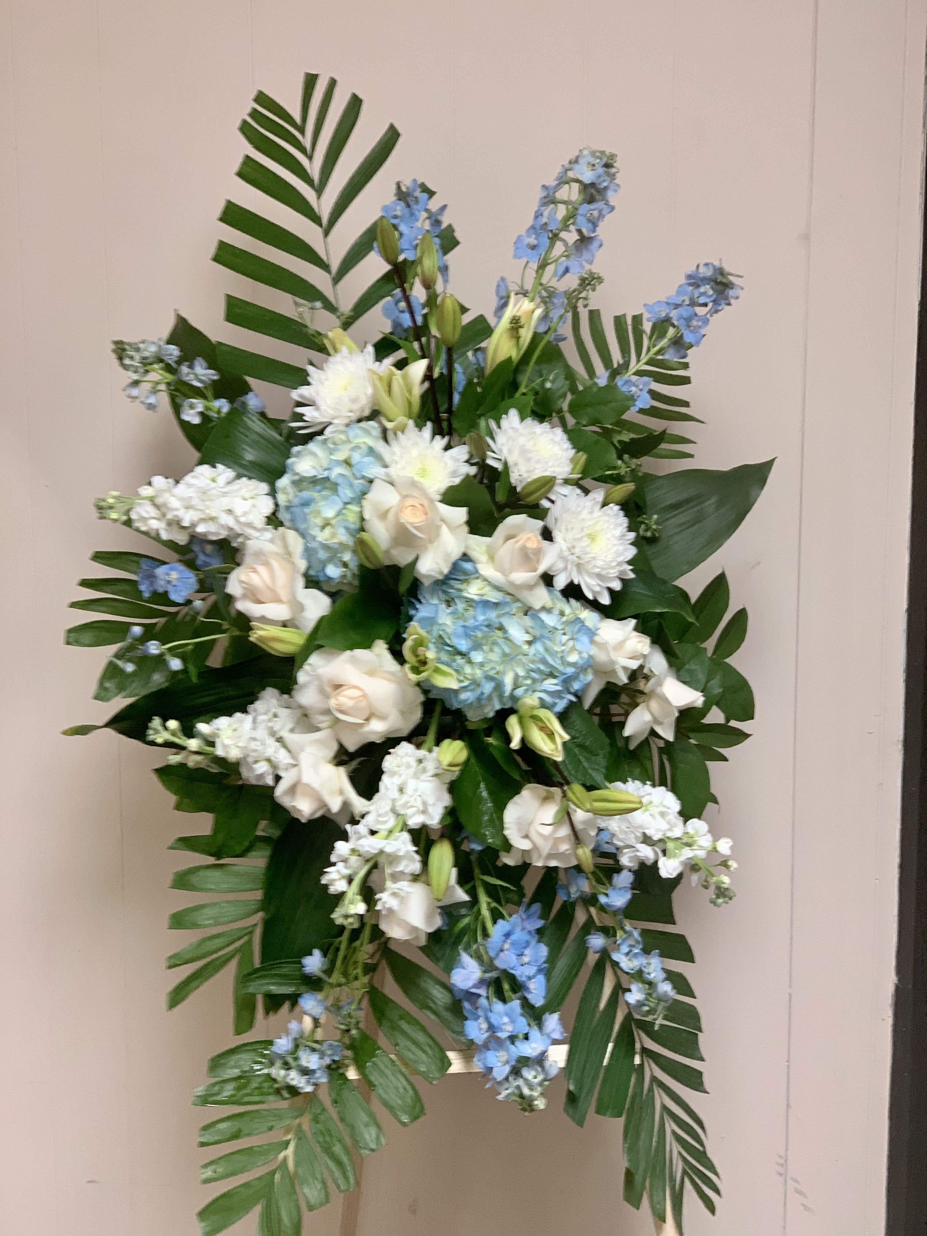 SYM23 - Ocean Breeze Spray - Tranquil blue and white flowers reminiscent of a soft ocean breeze offer comfort. The standing spray presented on an easel includes fragrant white lilies and tropical greenery. This standing spray includes blue hydrangea and delphinium, white roses and asiatic lilies, plus tropical ferns and leaves. Honor your loved one with this peaceful display of blooms, perfect for a funeral or memorial service. 