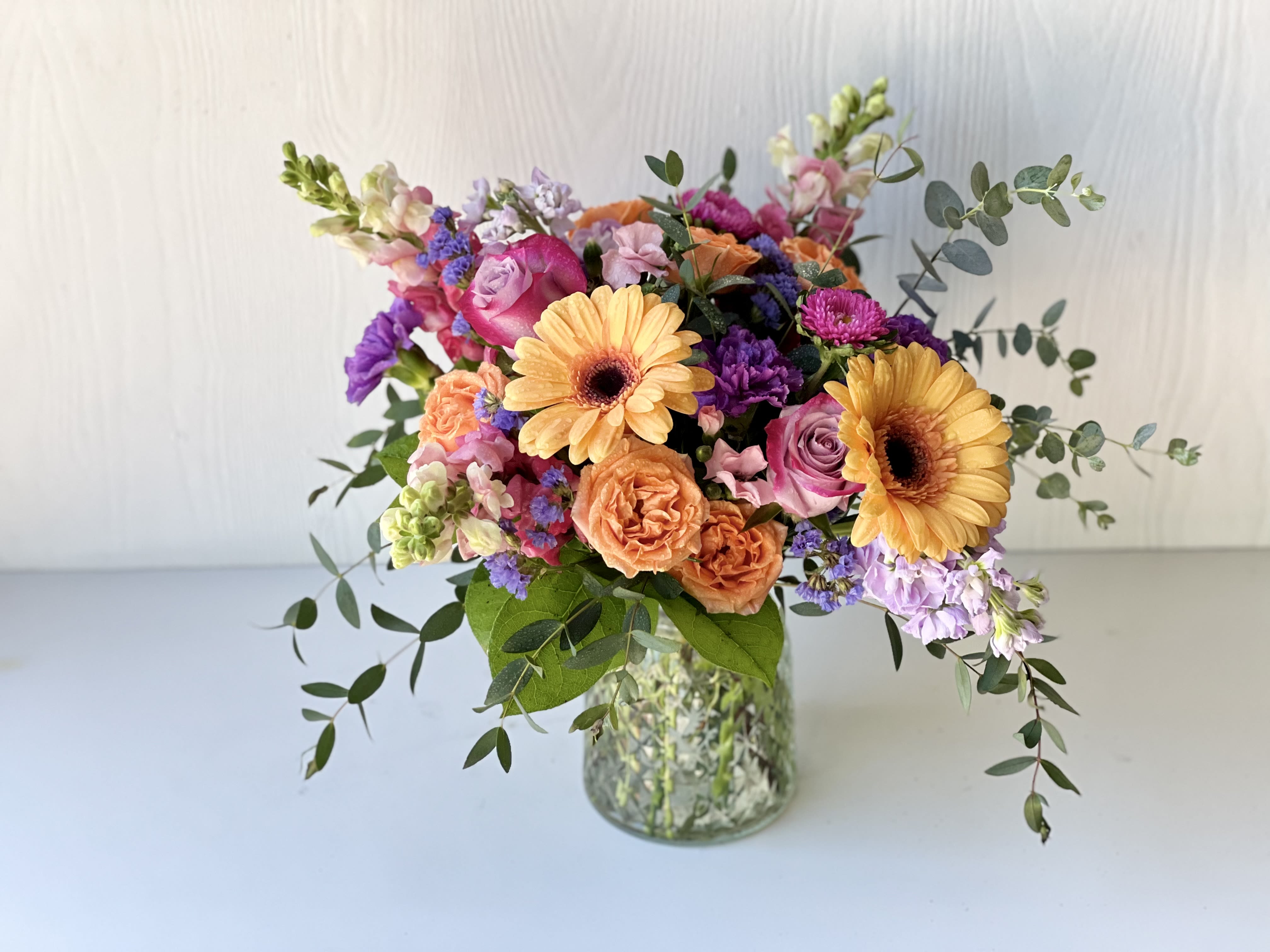 Mixed bouquet of orange gerbera daisies, pink roses, and purple flowers in a glass vase