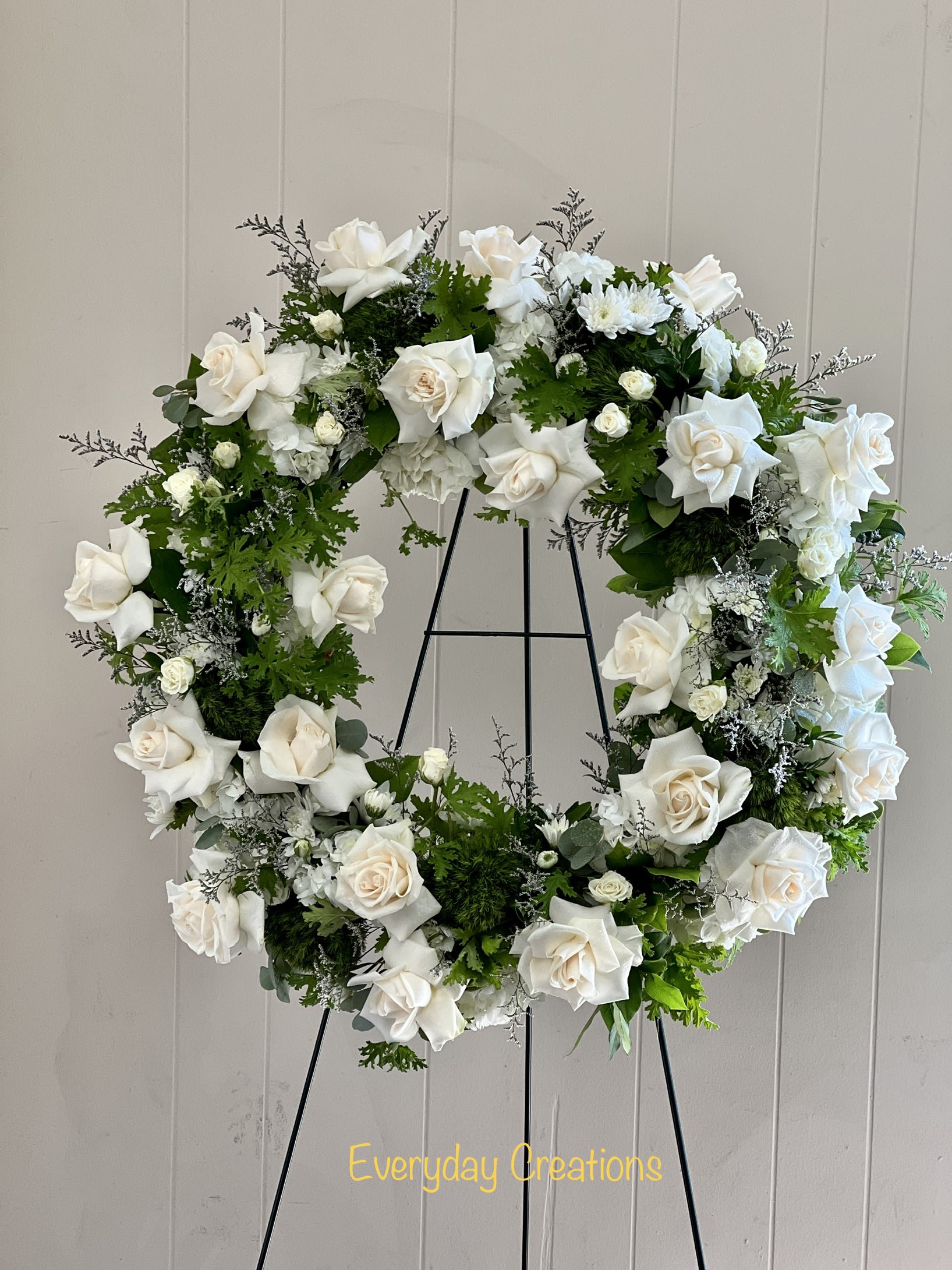 SYM20 - Sincere Condolences Wreath - A beautiful white and green tribute wreath that calms, this easel arrangement celebrates a full life and a peaceful passing. The Lasting Serenity standing spray features gorgeous flowers including roses, sprider mums, chrysanthemums and delicate grass woven throughout the wreath. This peaceful and elegant standing spray is delivered with a wire easel and is a perfect choice for a funeral, memorial service or any special occasion. 