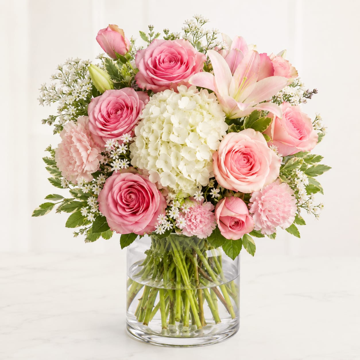 Kim’s Pink Elegance – Same Day Flower Delivery Houston - Soft blush roses and pale pink lilies form a rounded, romantic bouquet that reads like a gentle embrace-perfect as an anniversary arrangement or a "just because" gift to make someone feel seen. Pink, white and green tones combine here for a classic spring-ready look that also suits birthdays, Mother's Day, thank-you gestures, thinking-of-you moments, and love and romance occasions.  Tightly clustered pink roses and pale pink lilies are balanced by creamy white and blush hydrangea blooms, small white filler flowers, soft pink carnations, tulips, glossy foliage and visible stems arranged in a clear glass vase. The composition is full and polished rather than loose, with a compact, upright silhouette about the size of a traditional centerpiece; the clear vase gives a fresh, clean presentation and lets the natural stems show through.  This is the kind of bouquet that will make the person you're thinking of feel truly celebrated-whether you're saying happy birthday, marking an anniversary, or sending a tender thank you. Ready for same-day flower delivery in Houston, it's arranged from fresh, top-quality blooms and arrives ready to display; change the water and recut stems every few days to help prolong its beauty.