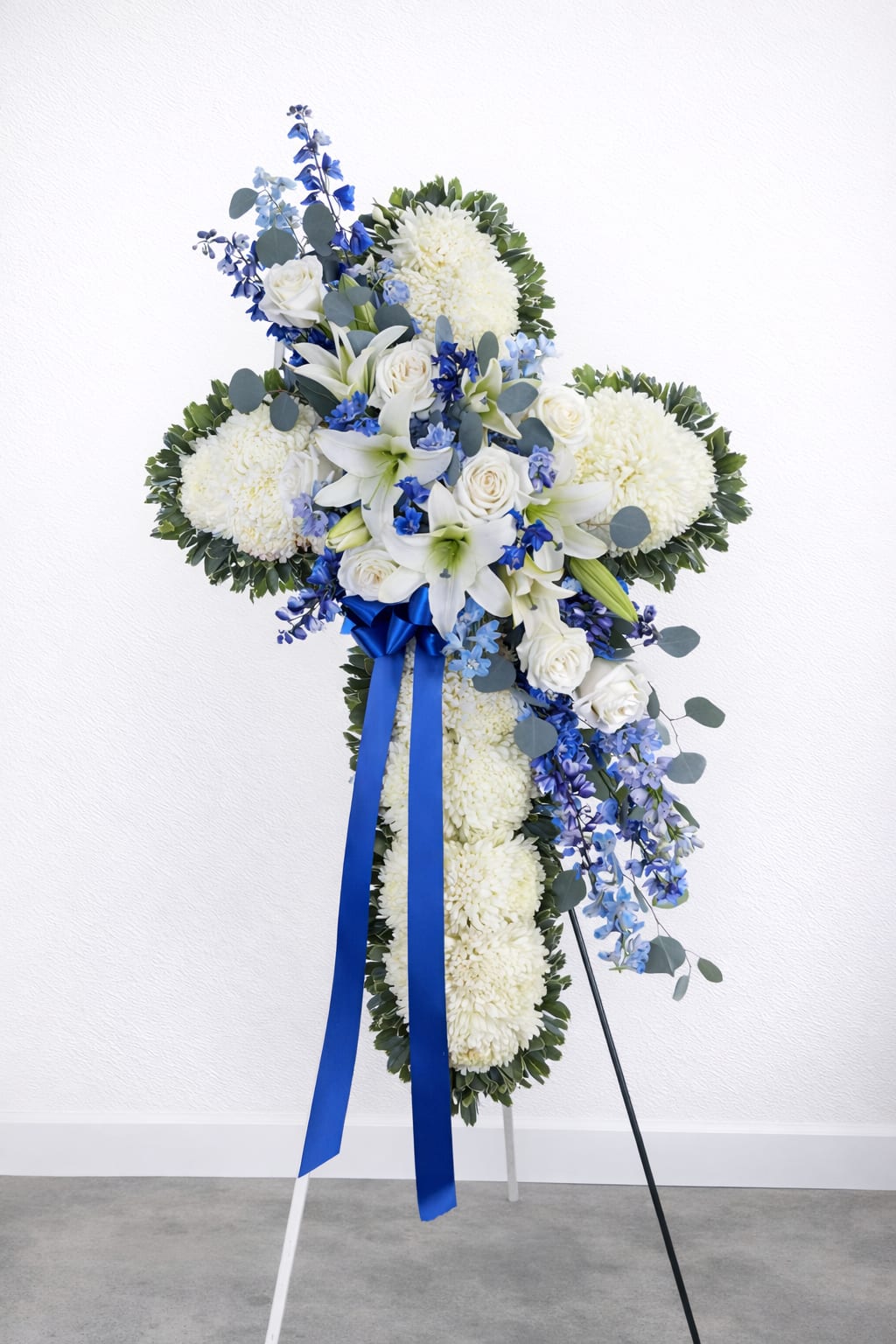 Deepest Faith Cross - Standing cross arrangement with white football mums, roses and Oriental lilies; blue delphinium; accented with silver dollar eucalyptus, assorted greenery and blue ribbon