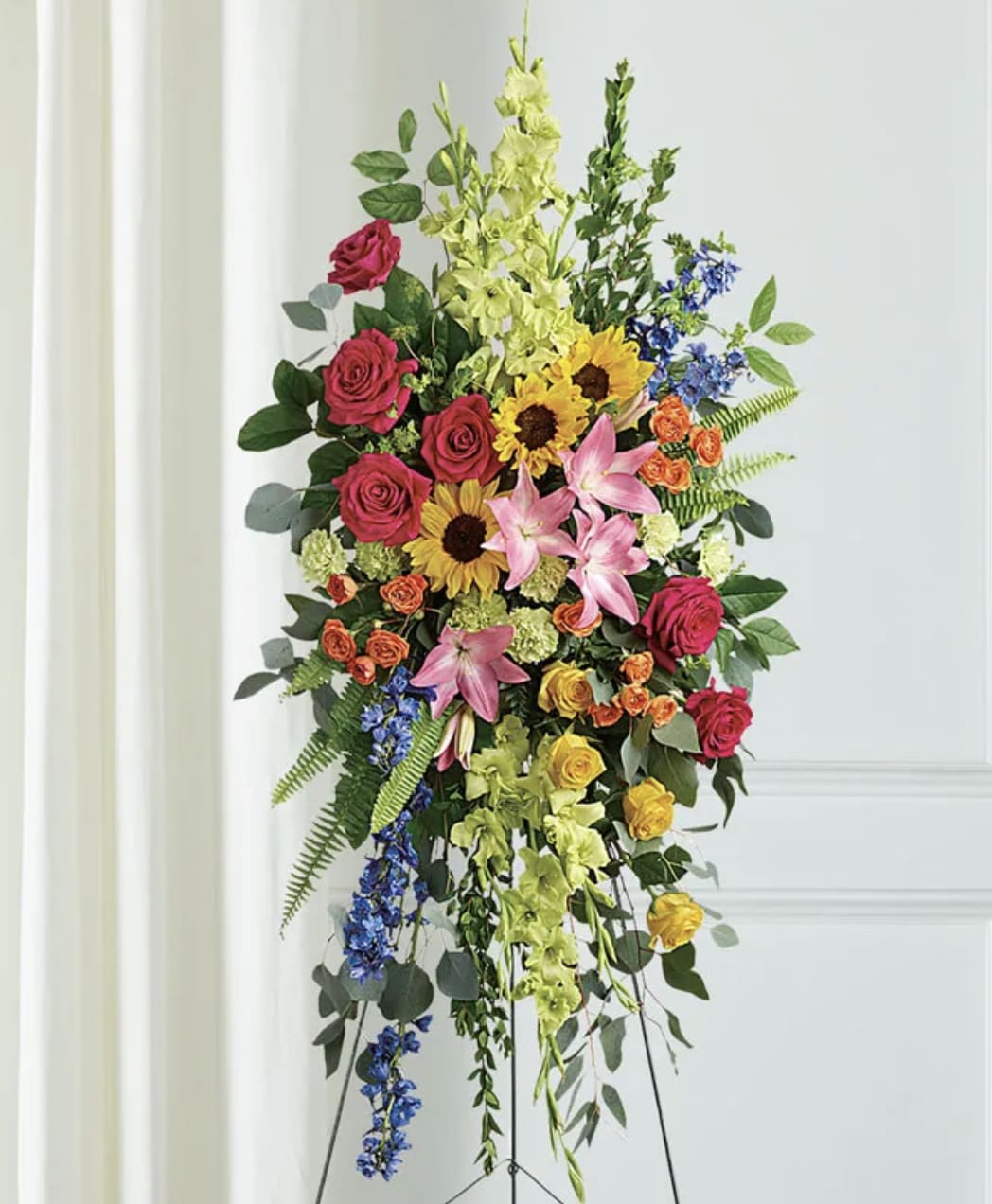 Tall floral standing spray with roses, lilies, sunflowers, and blue accents