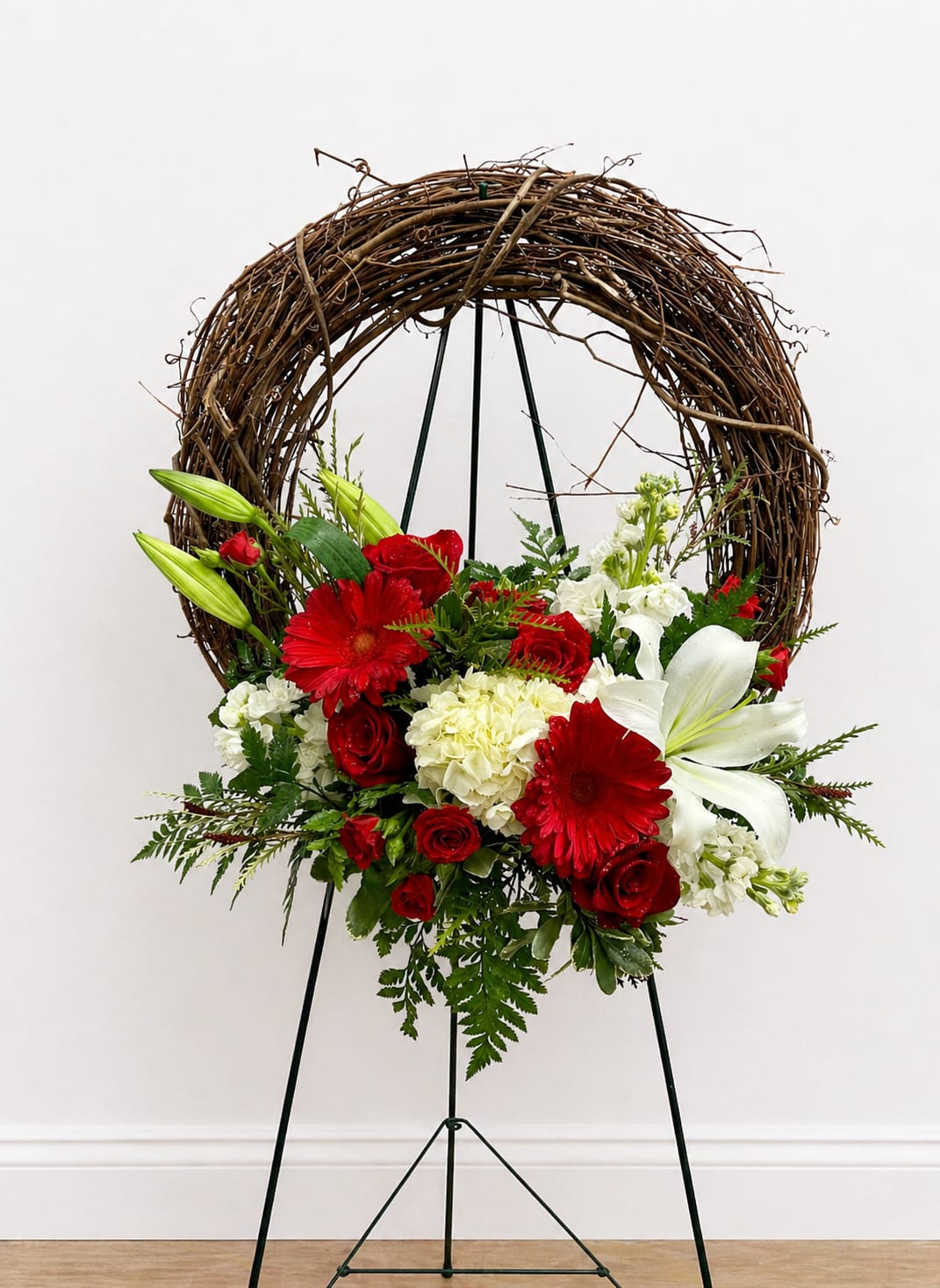 Vine Wreath - This is a floral arrangement designed on a circular grapevine wreath mounted on a black metal easel. The wreath itself is made of intertwined natural brown vines, giving it a rustic, organic look.  At the bottom center of the wreath sits a lush, asymmetrical flower arrangement. The color palette is primarily red, white, and green, creating a bold and elegant contrast. Key flowers include:  Bright red roses and red gerbera daisies, which stand out as the focal points White lilies with open petals, adding a soft, graceful touch Cream or white carnations, giving fullness and texture A few unopened lily buds for variation and height  The flowers are surrounded by rich greenery—ferns and leafy stems—that add depth and a natural flow to the design.  Overall, the piece feels like a decorative wreath arrangement suitable for occasions such as memorials, holidays, or formal events, combining rustic structure with a classic, vibrant floral display.