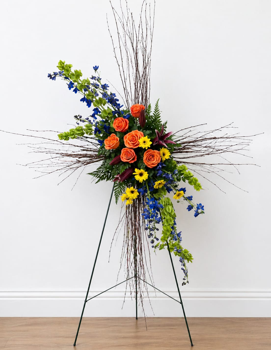 Colorful Grapevine Cross - This is a modern, artistic standing floral arrangement displayed on a metal easel, built around a striking cross-like framework made of thin, natural branches.  At the center is a compact, vibrant cluster of flowers, featuring:  Bright orange roses as the main focal point Small yellow daisy-like blooms adding cheerful contrast Deep blue delphinium (or similar flowers) bringing bold color depth Light green accent flowers that add freshness and texture A few dark burgundy lilies for richness and variety  Surrounding the center, long, thin branches extend outward horizontally and vertically, forming a loose cross or starburst shape. Some branches also cascade downward, giving the design movement and a slightly dramatic, flowing effect.  The greenery—ferns and leafy foliage—fills in the base, grounding the bright colors while keeping the overall look balanced.  Overall, the arrangement feels contemporary and expressive, with a mix of structured shape (the cross-like branch design) and organic flow. It would be suitable for events like memorials, celebrations of life, or even artistic displays where a bold, modern floral statement is desired.