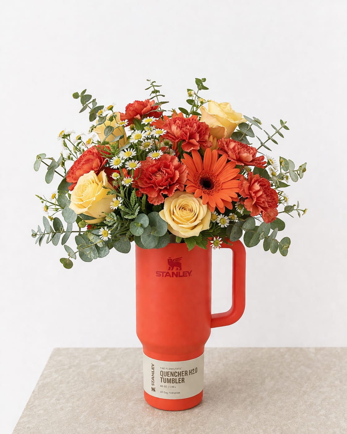 Sunset Stanley Floral Arrangement - Bright, cheerful, and full of warmth, this stunning arrangement is designed in a vibrant orange Stanley tumbler for a gift that’s both beautiful and practical. Featuring a lively mix of golden yellow roses, bold orange gerbera daisies, and textured carnations, this design is accented with delicate white daisies and fresh eucalyptus for a natural, garden-inspired feel.  The warm color palette creates a radiant, feel-good vibe—perfect for birthdays, celebrations, or simply brightening someone’s day. Once the flowers are enjoyed, the reusable Stanley tumbler becomes a lasting keepsake for everyday use.  Handcrafted with care, each arrangement is uniquely styled to create a full, balanced look that highlights the beauty of every bloom.